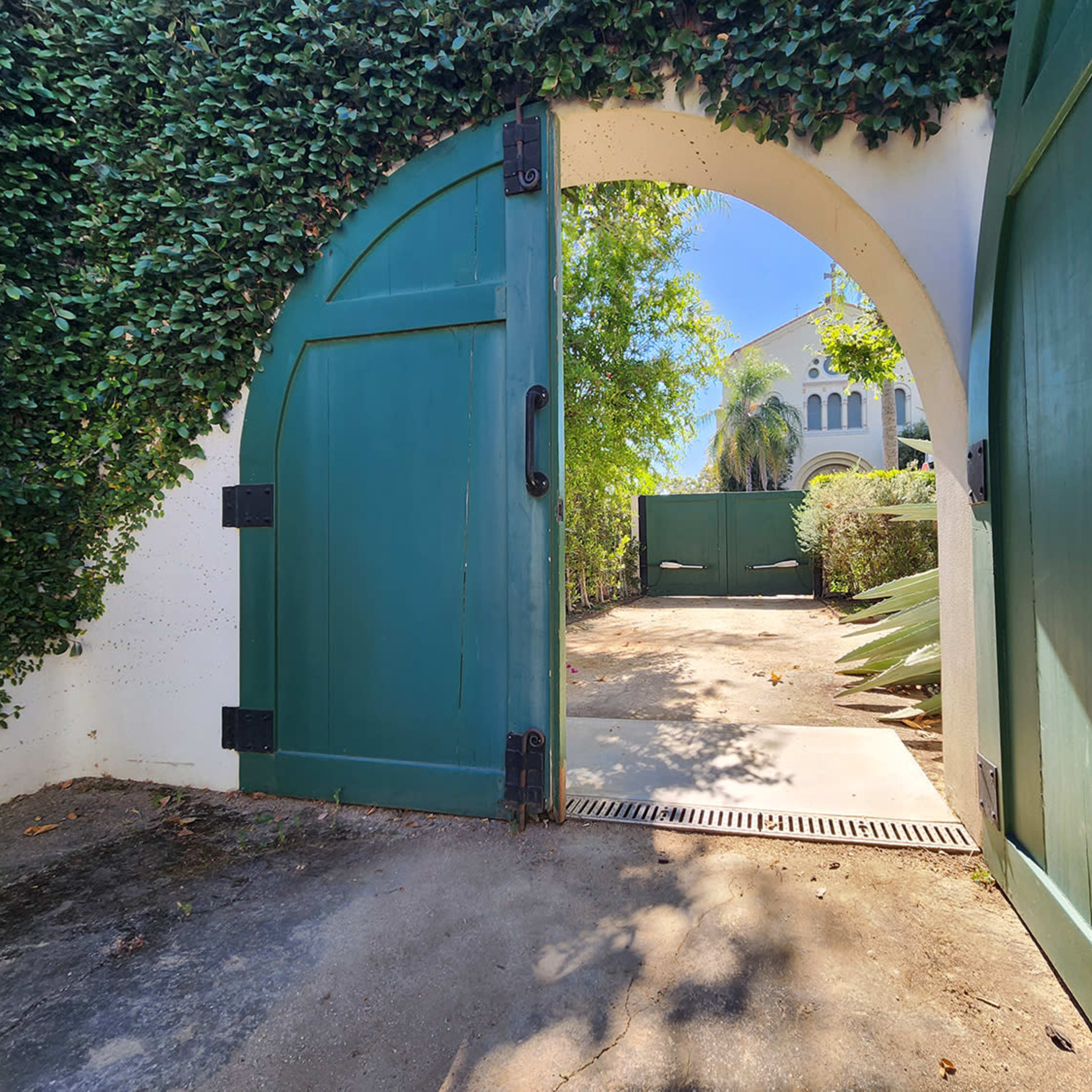 An arched green gate opens to a sunny pathway surrounded by lush foliage and a white wall.