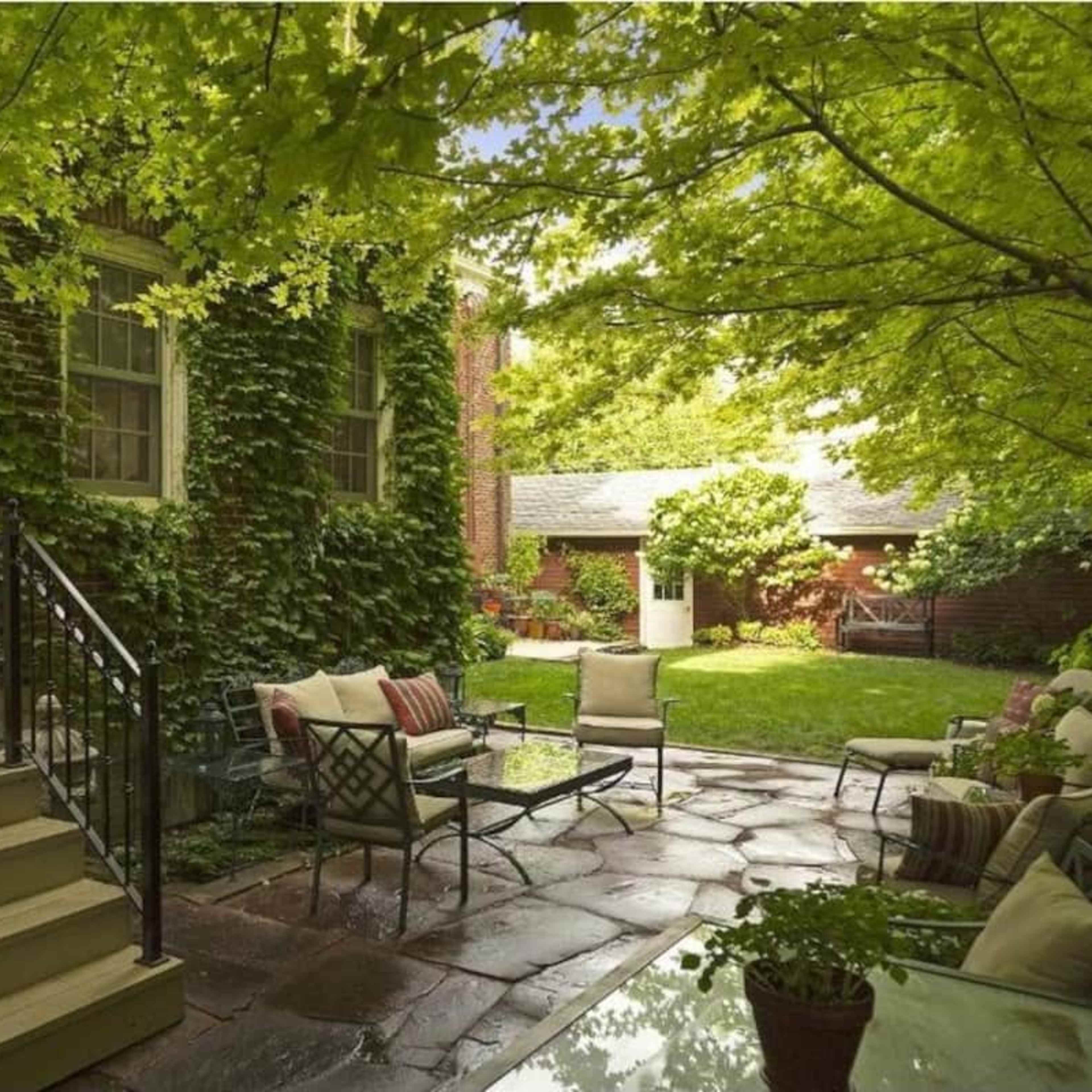 A stone patio with seating and a staircase leads to a brick house surrounded by greenery and plants.