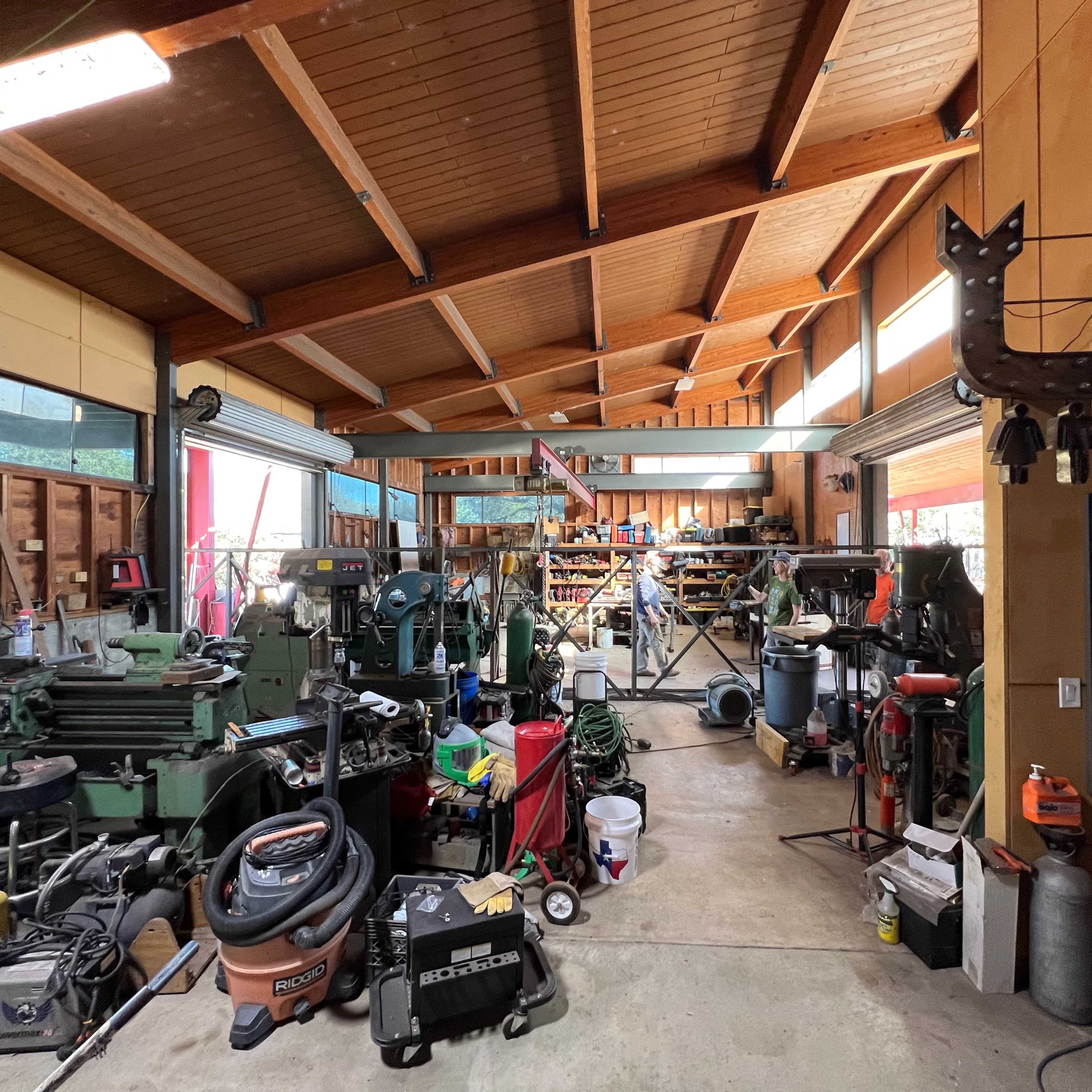 A large workshop filled with various tools, machines, and equipment, with a high wooden ceiling and ample natural light coming through large windows.