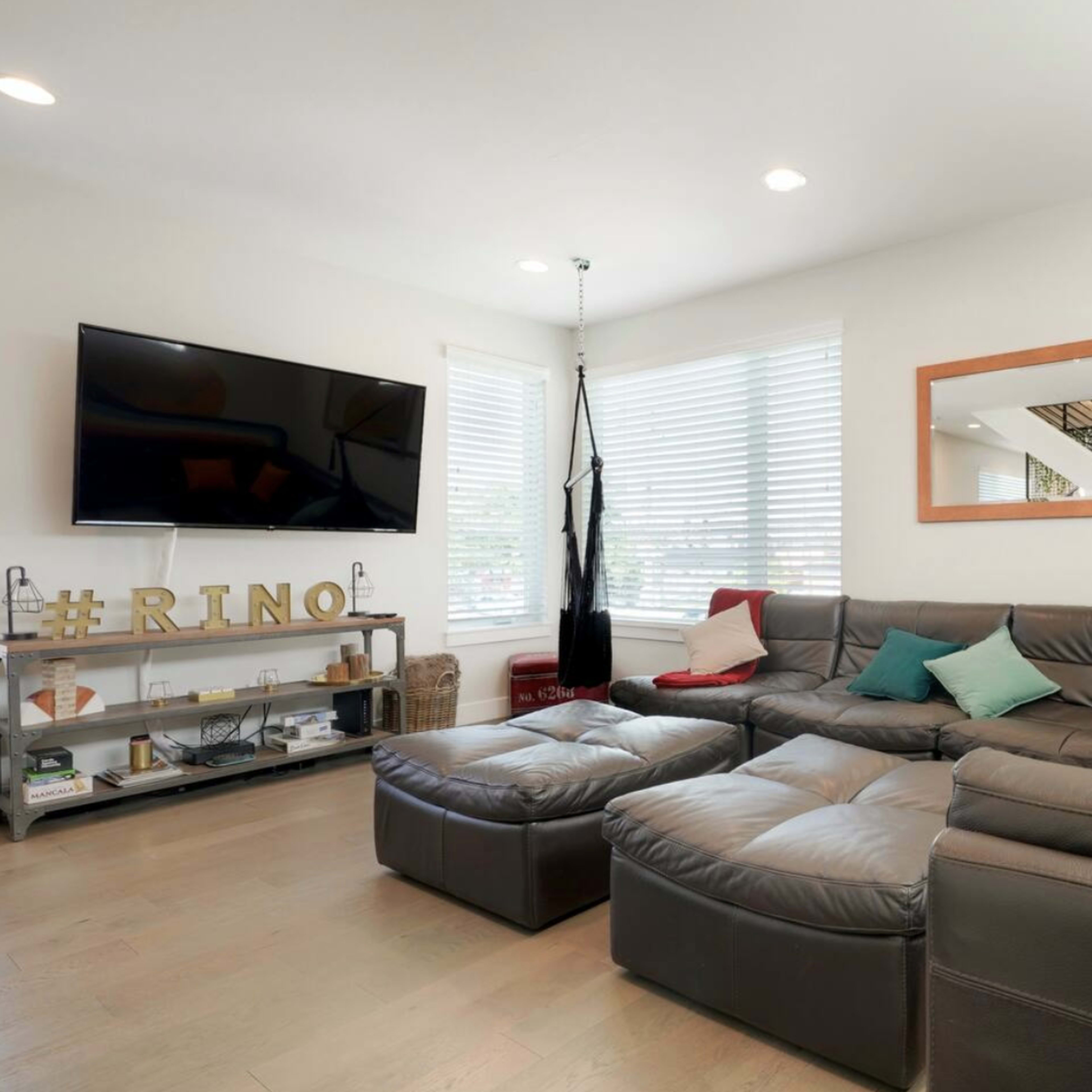 A modern living room features a large sectional sofa with colorful pillows, a flat-screen TV mounted on the wall, and a shelf displaying decorative items.
