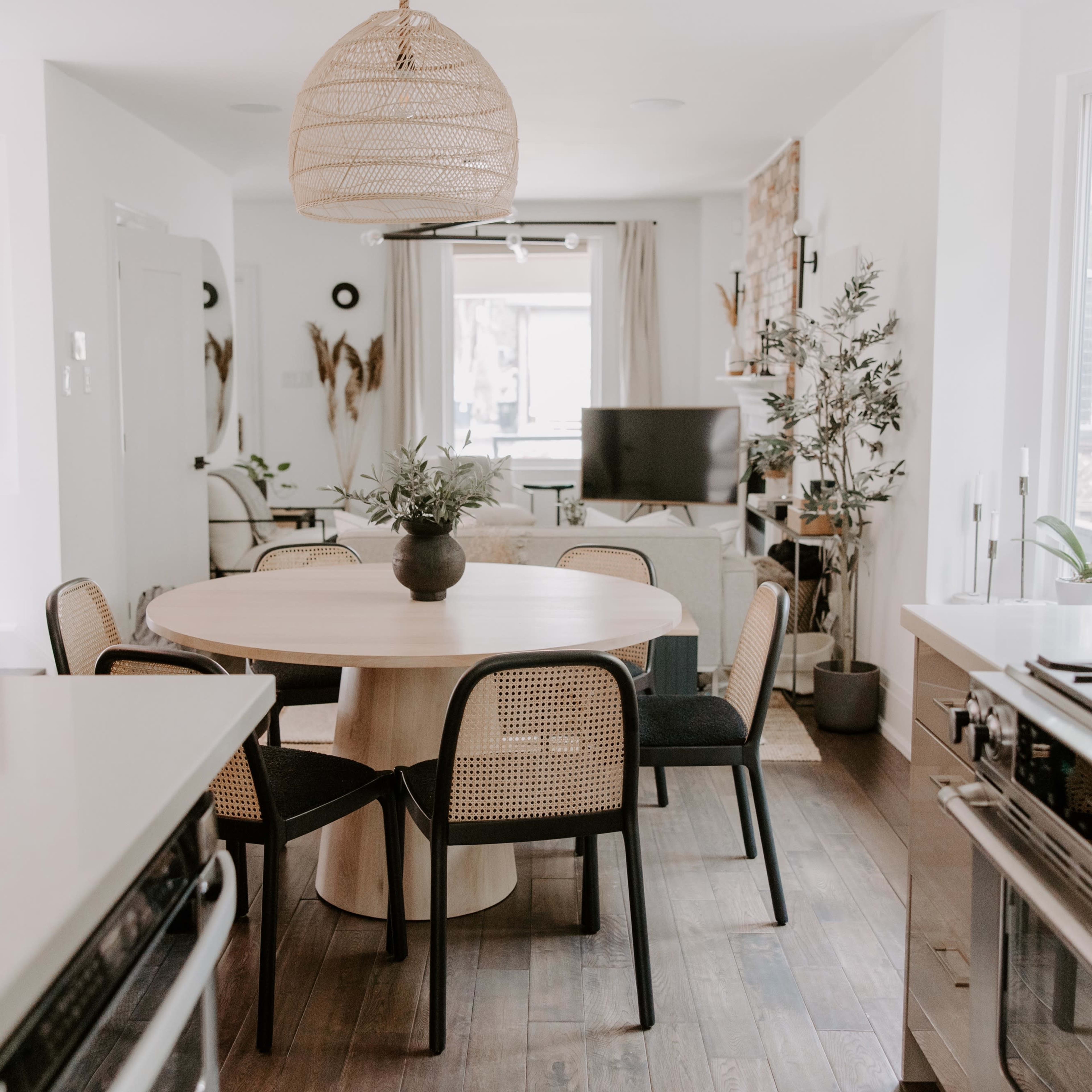 A dining area features a round wooden table surrounded by four chairs, with a modern kitchen and living space visible in the background.