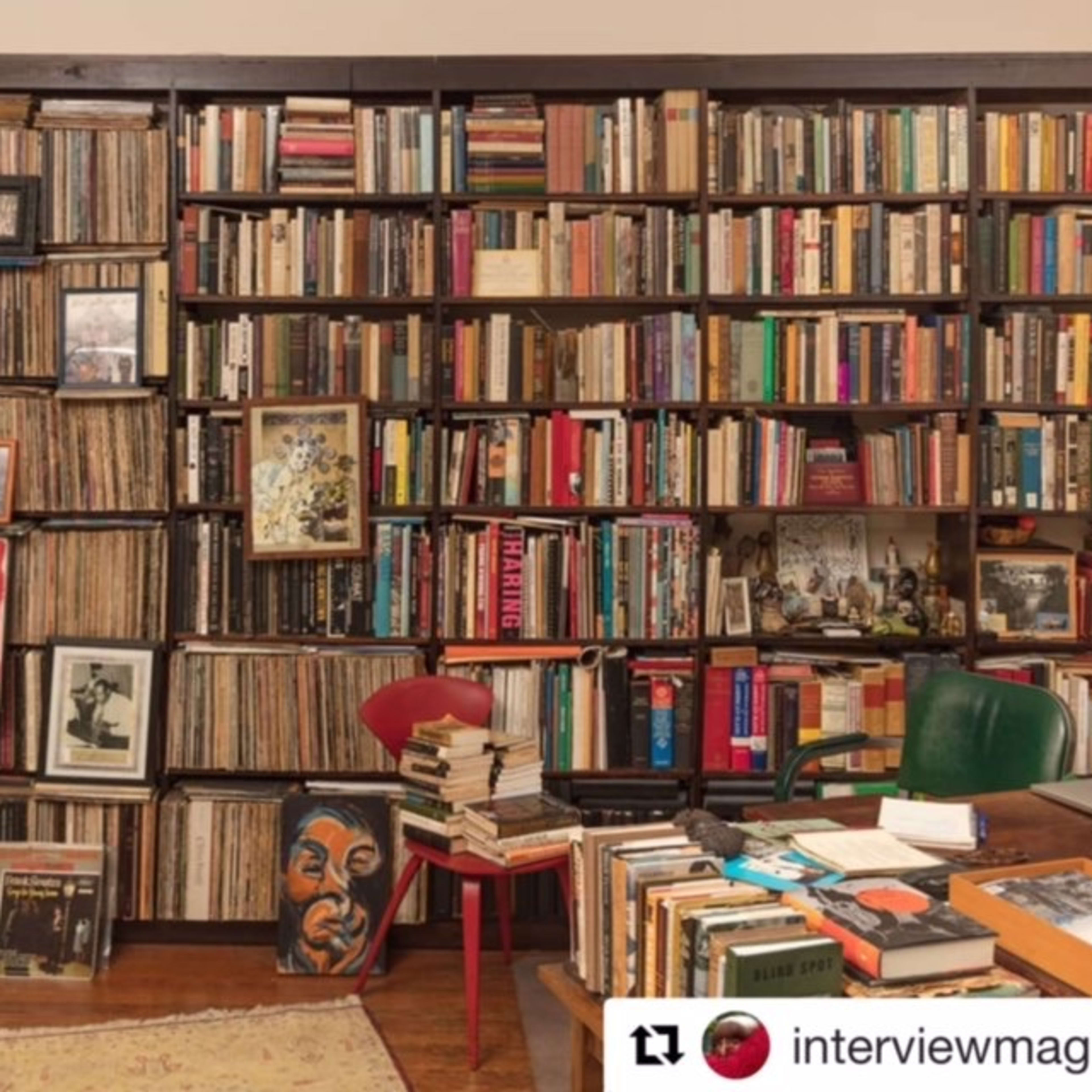 A wooden bookshelf filled with various books and vinyl records, flanked by a small table stacked with more books and two chairs.