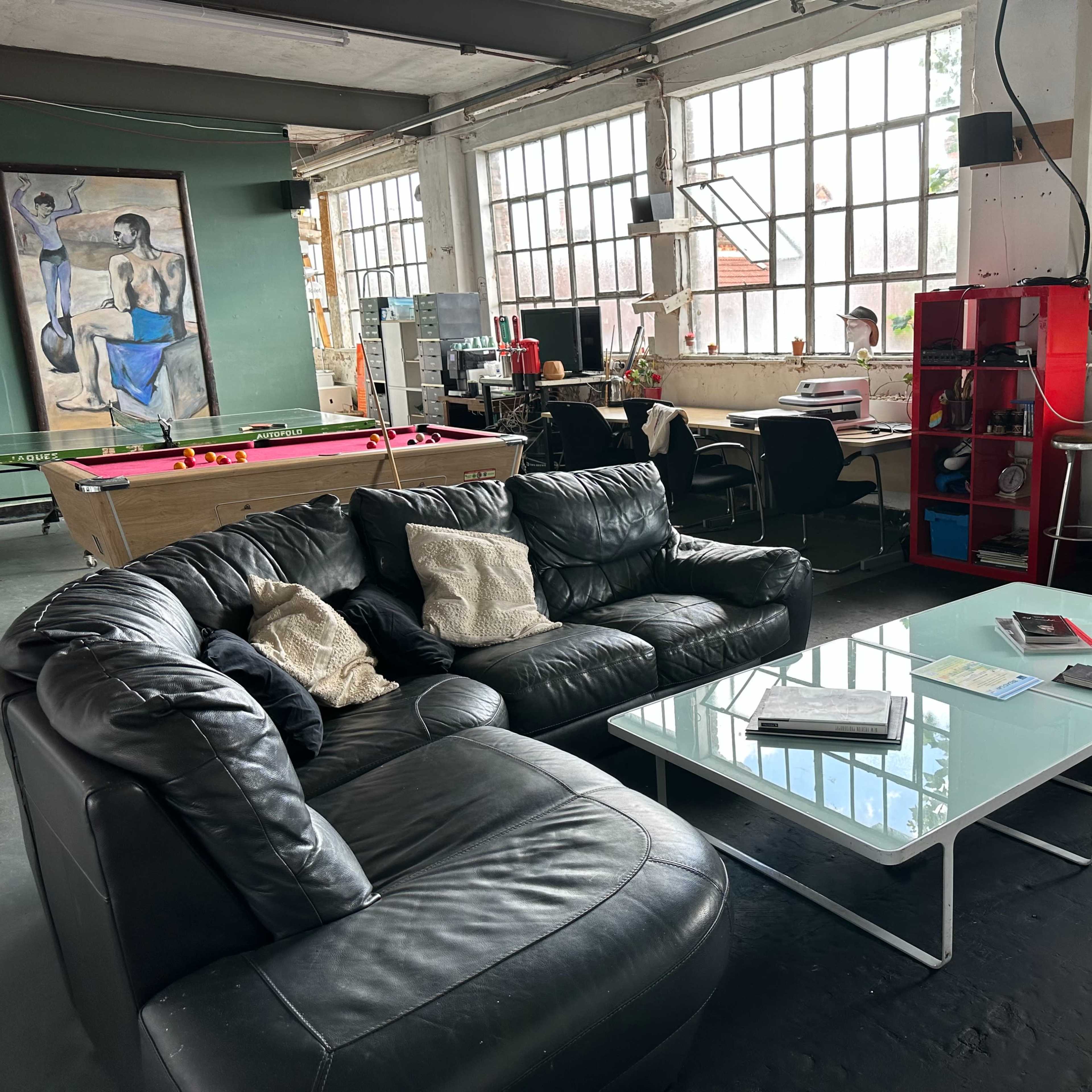A spacious industrial-inspired room with large windows, featuring a black leather sofa, a glass coffee table, and workstations in the background.