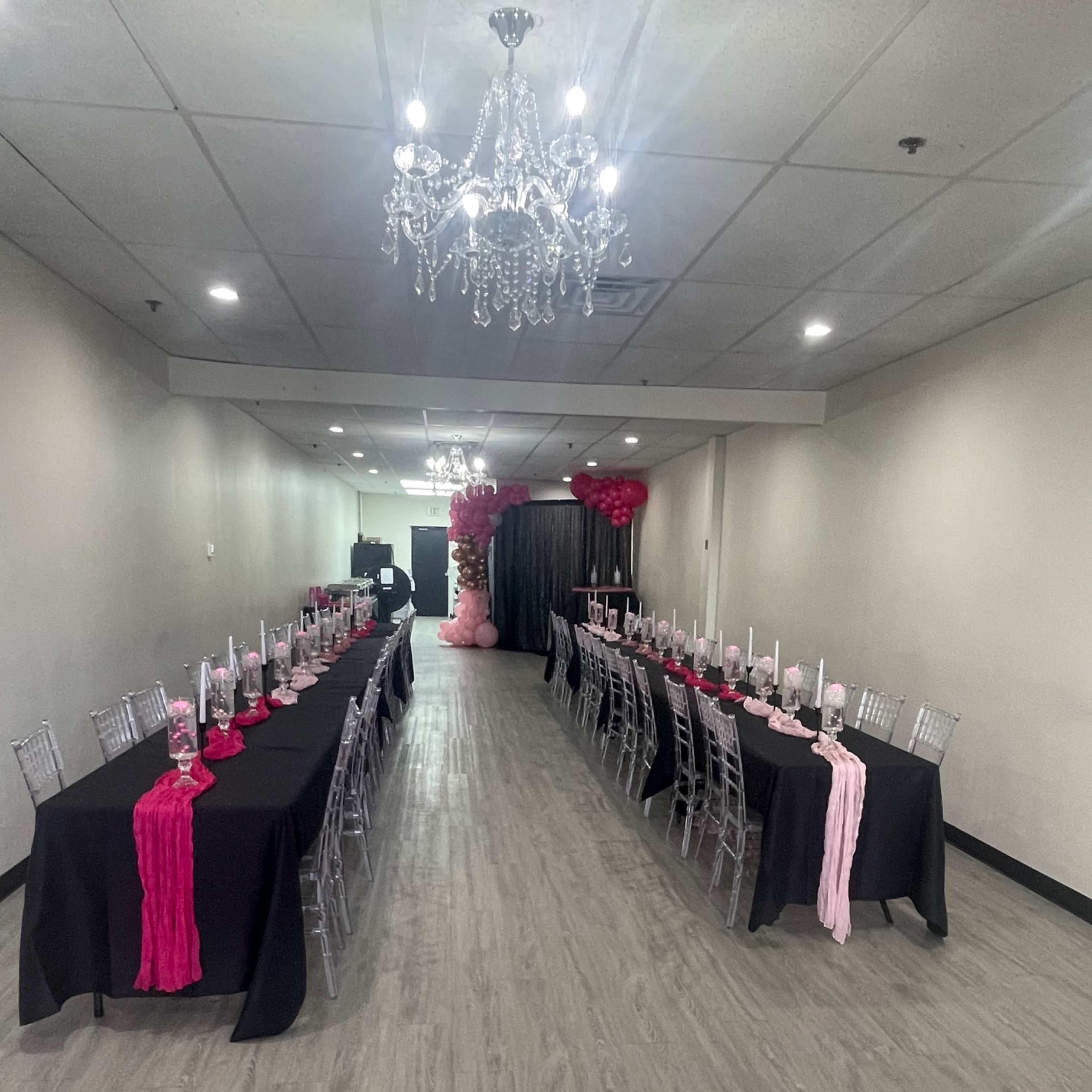 A long banquet hall features two rows of elegantly set tables with black tablecloths and decorative pink accents.