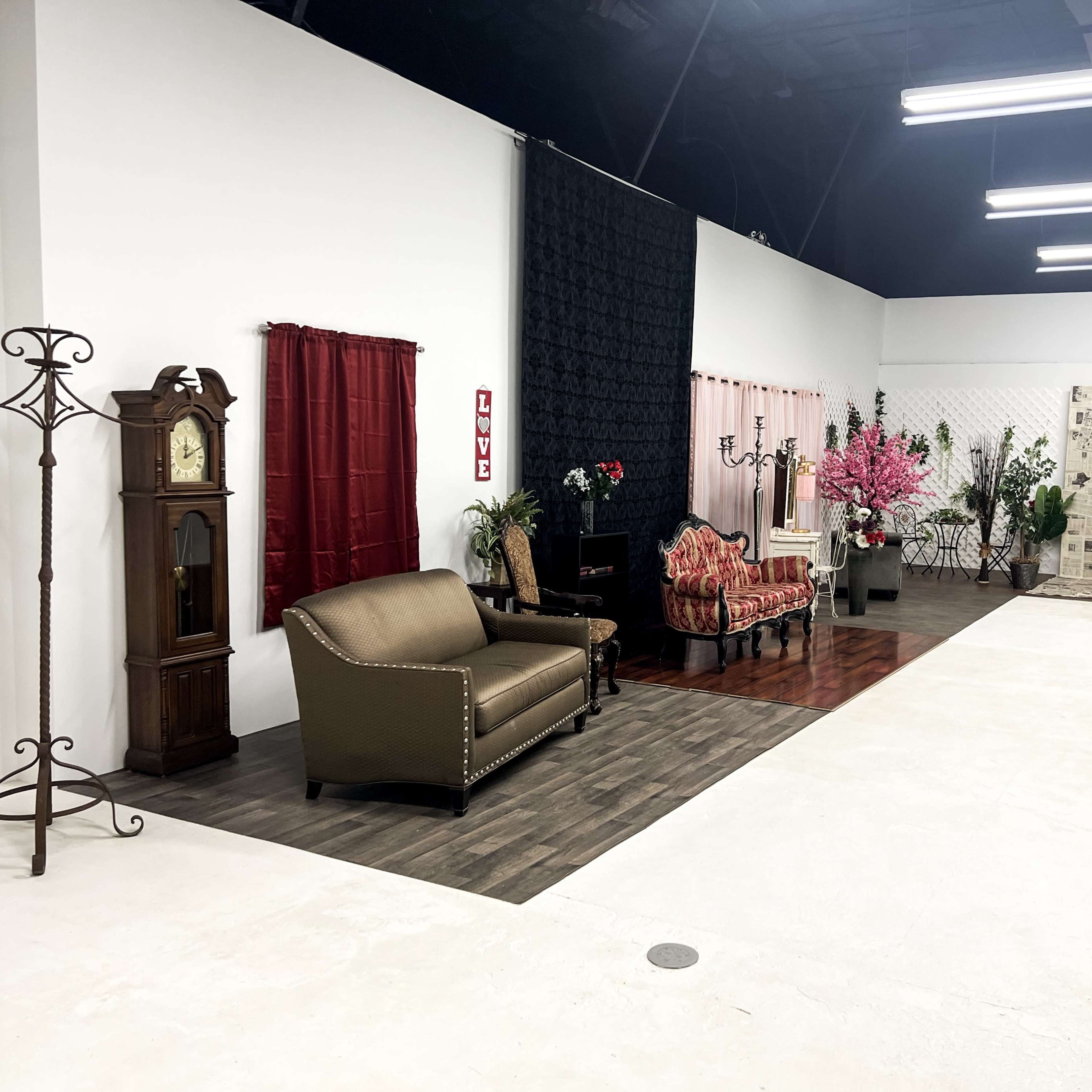 An interior studio space featuring various seating arrangements, decorative curtains, a grandfather clock, and ornamental plants.