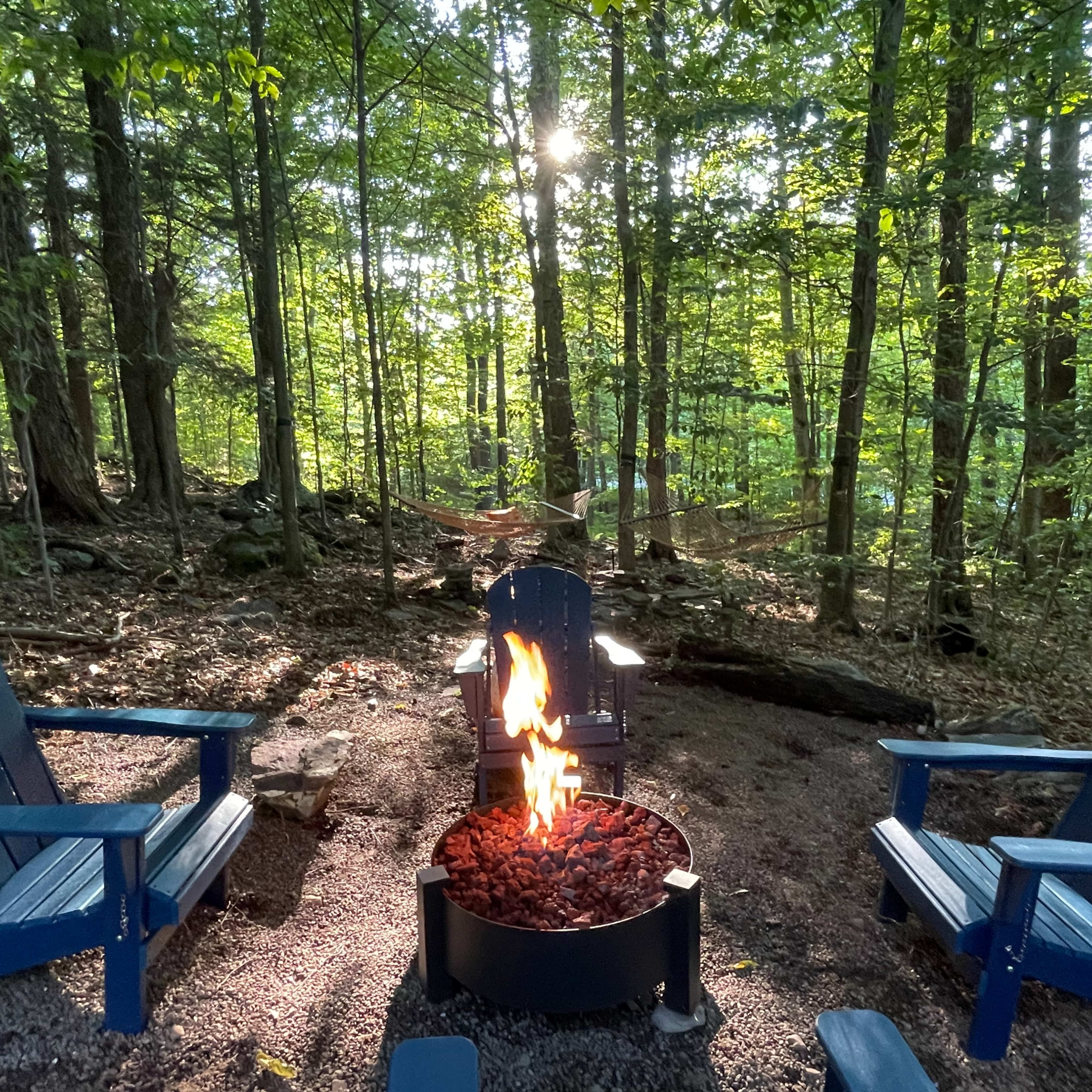 A circular fire pit with a flame surrounded by blue adirondack chairs sits in a forested area with trees and a hammock in the background.