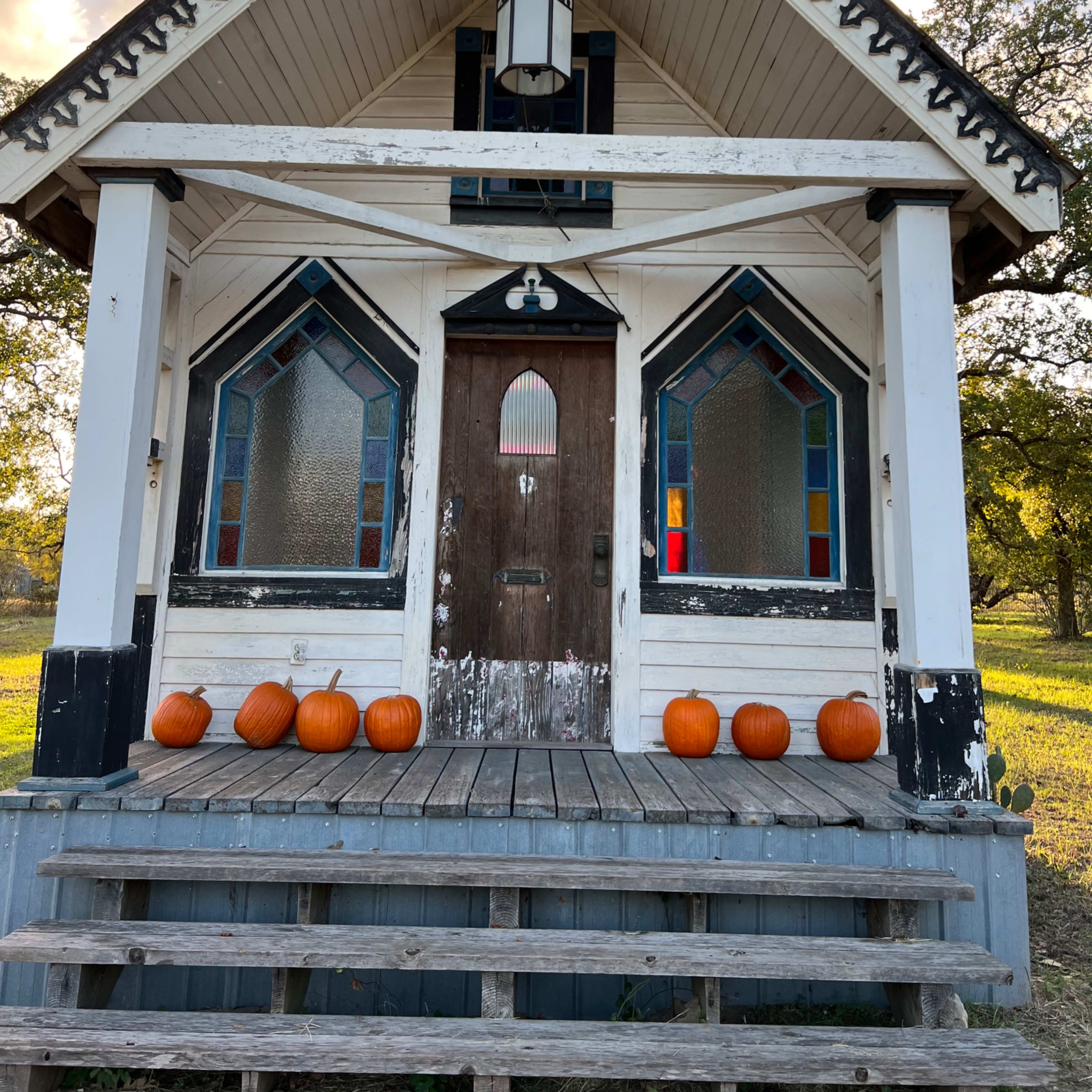 A small, decorative house with stained glass windows and pumpkins lined up on the front steps.