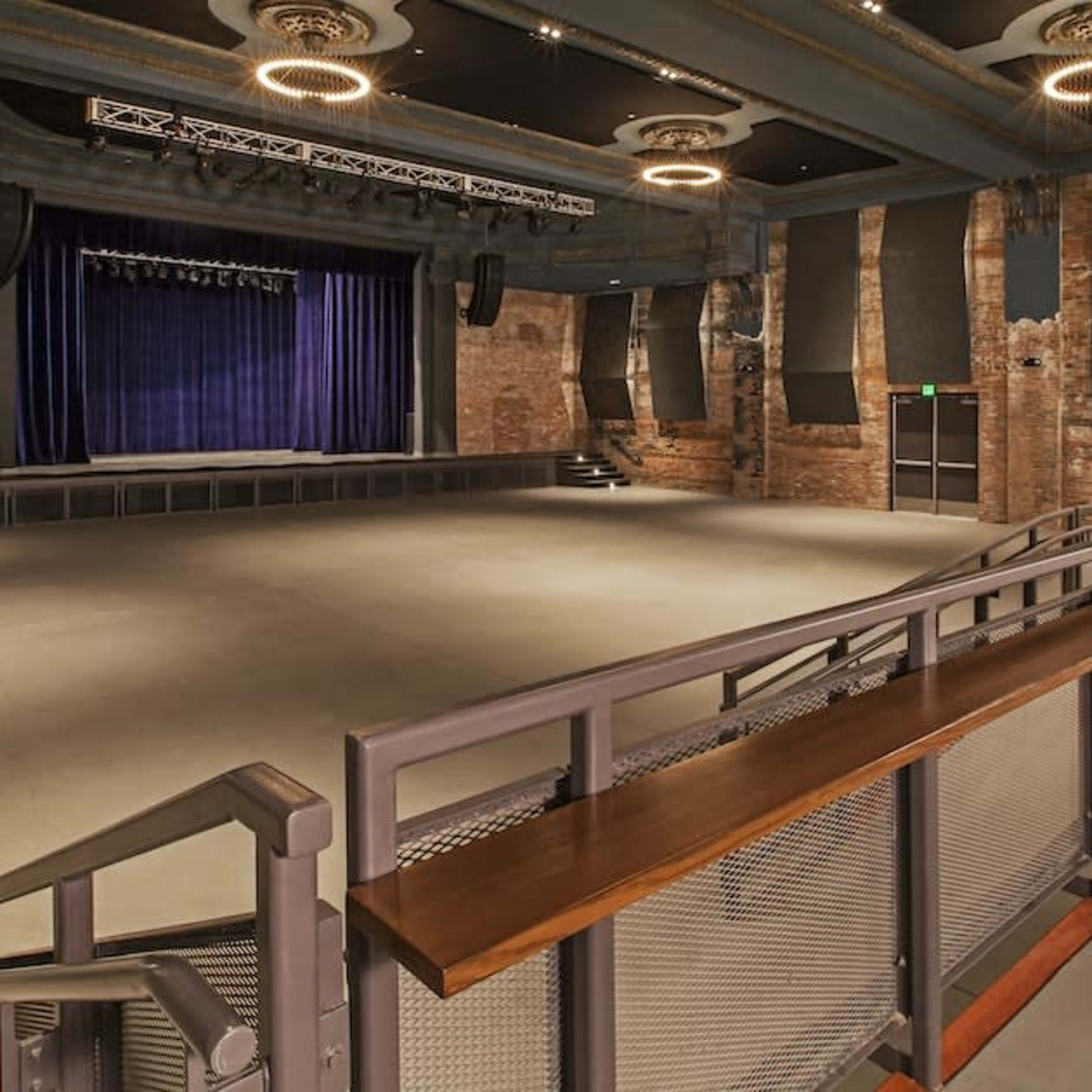 An empty auditorium with a wooden railing overlooking a stage surrounded by exposed brick walls and spotlights.