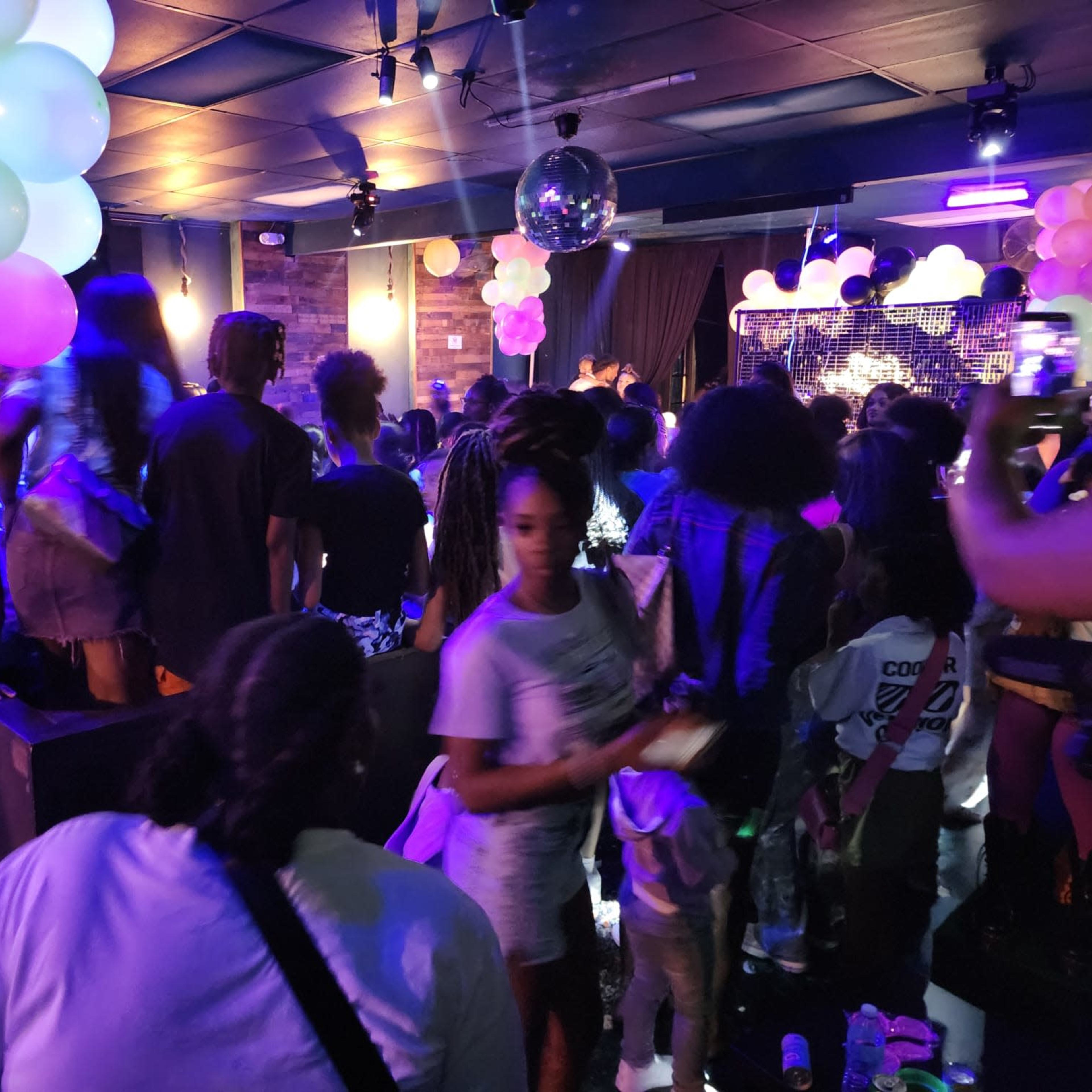 A crowded party scene features colorful balloons and a disco ball, with people socializing and dancing under dim lighting.