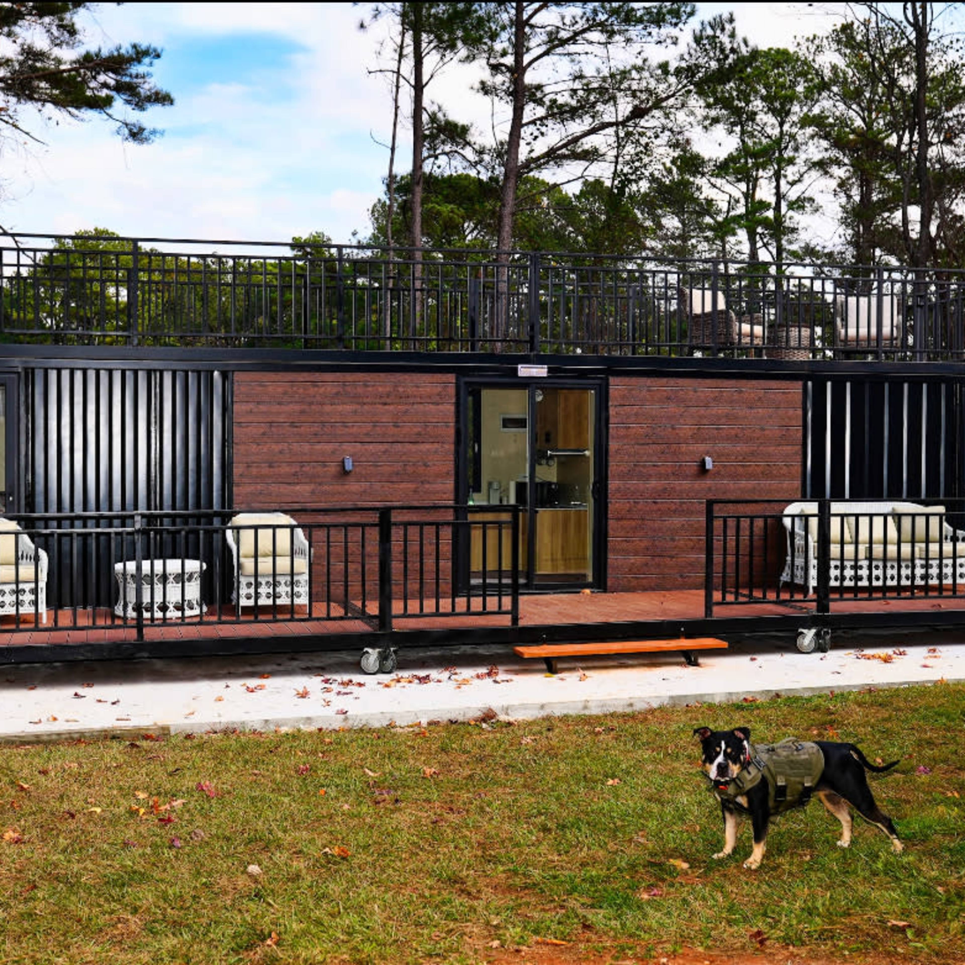 A modern, two-story shipping container home features a deck and seating area in front, with a dog in the foreground.