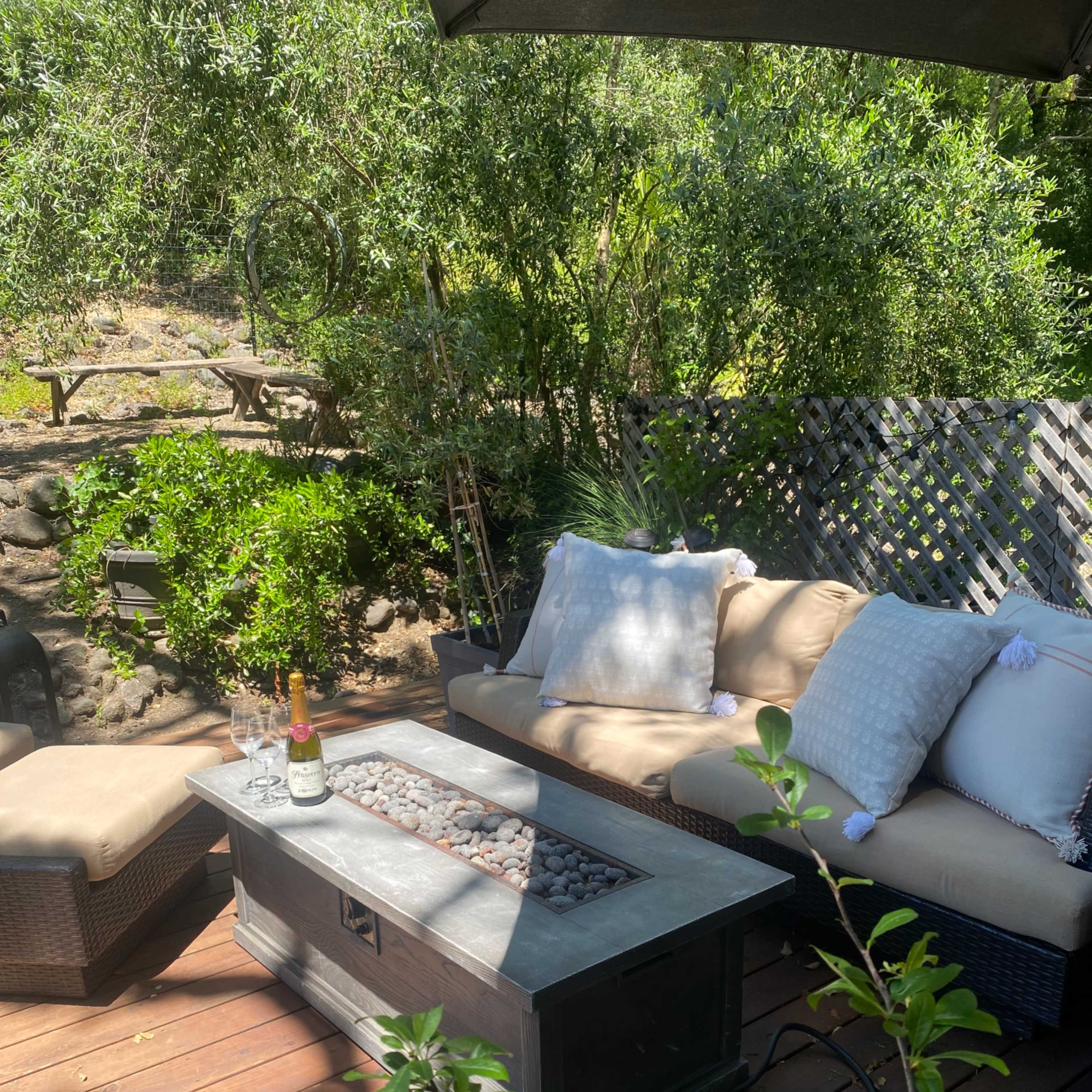 A patio area features a sectional couch with plush pillows, a stone fire table, and two glasses beside a bottle of champagne, surrounded by greenery.