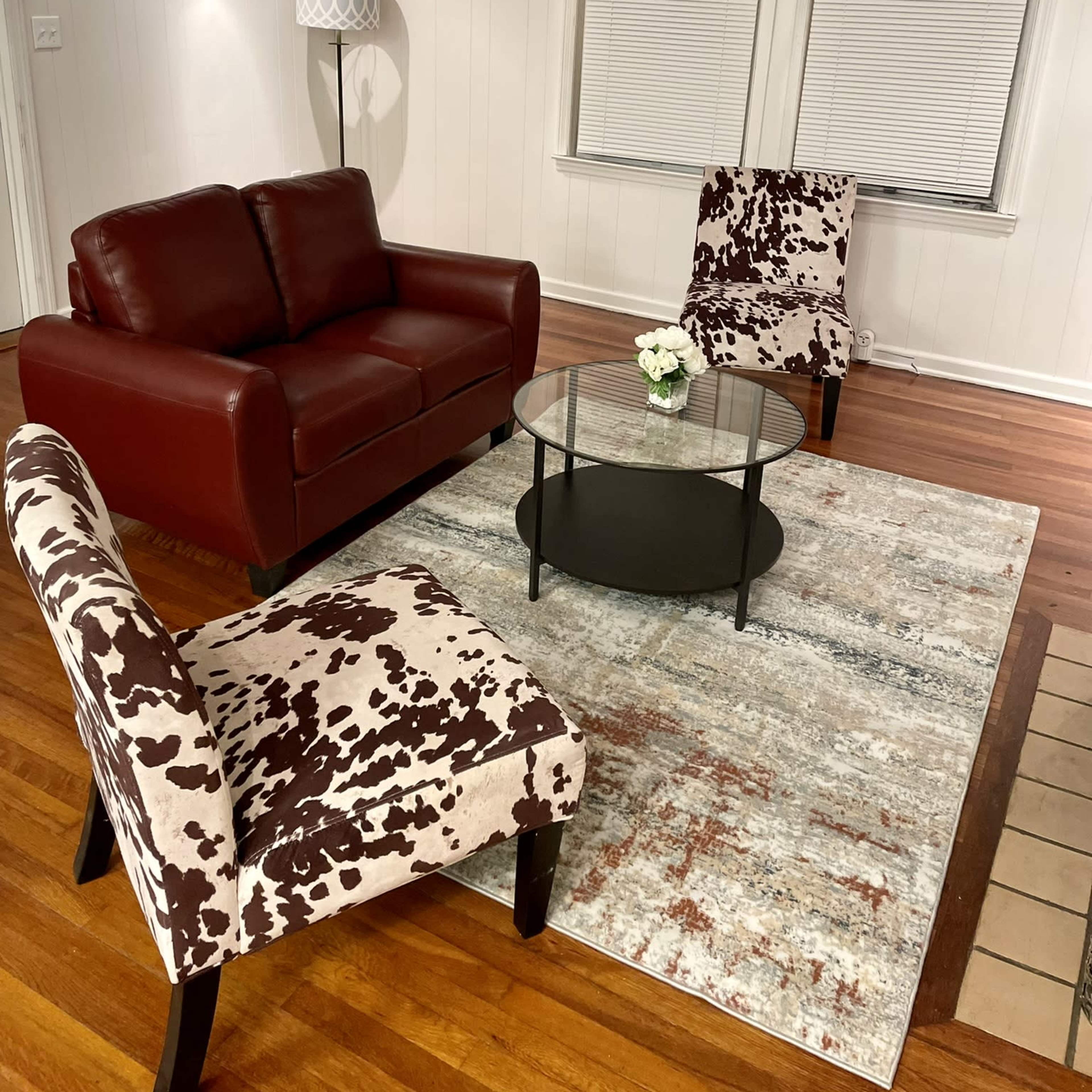 A living room with a red leather sofa, two patterned chairs, a glass coffee table on a rug, and a floral wall art piece.