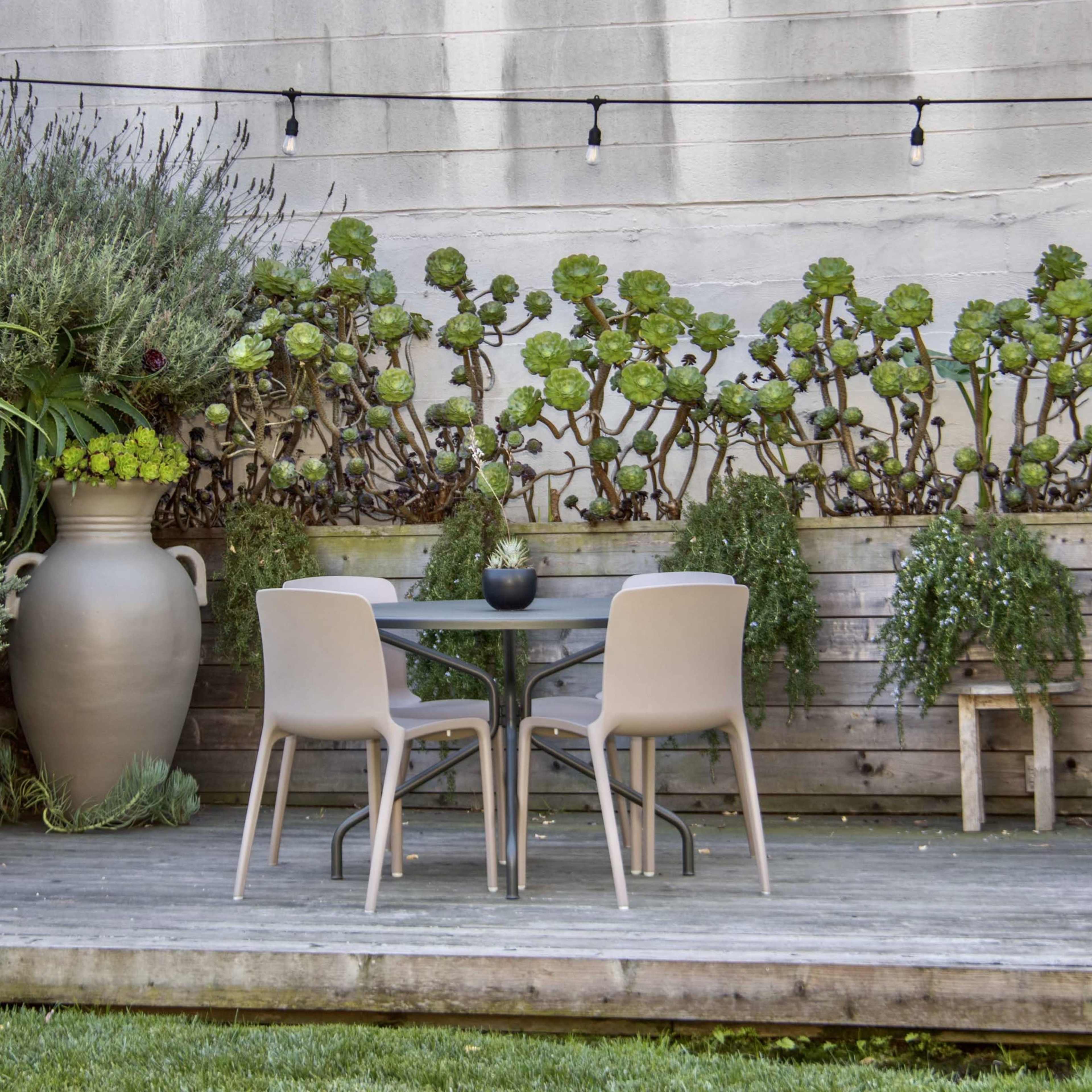 A small wooden deck features a round table and two chairs surrounded by potted plants and a large decorative vase.