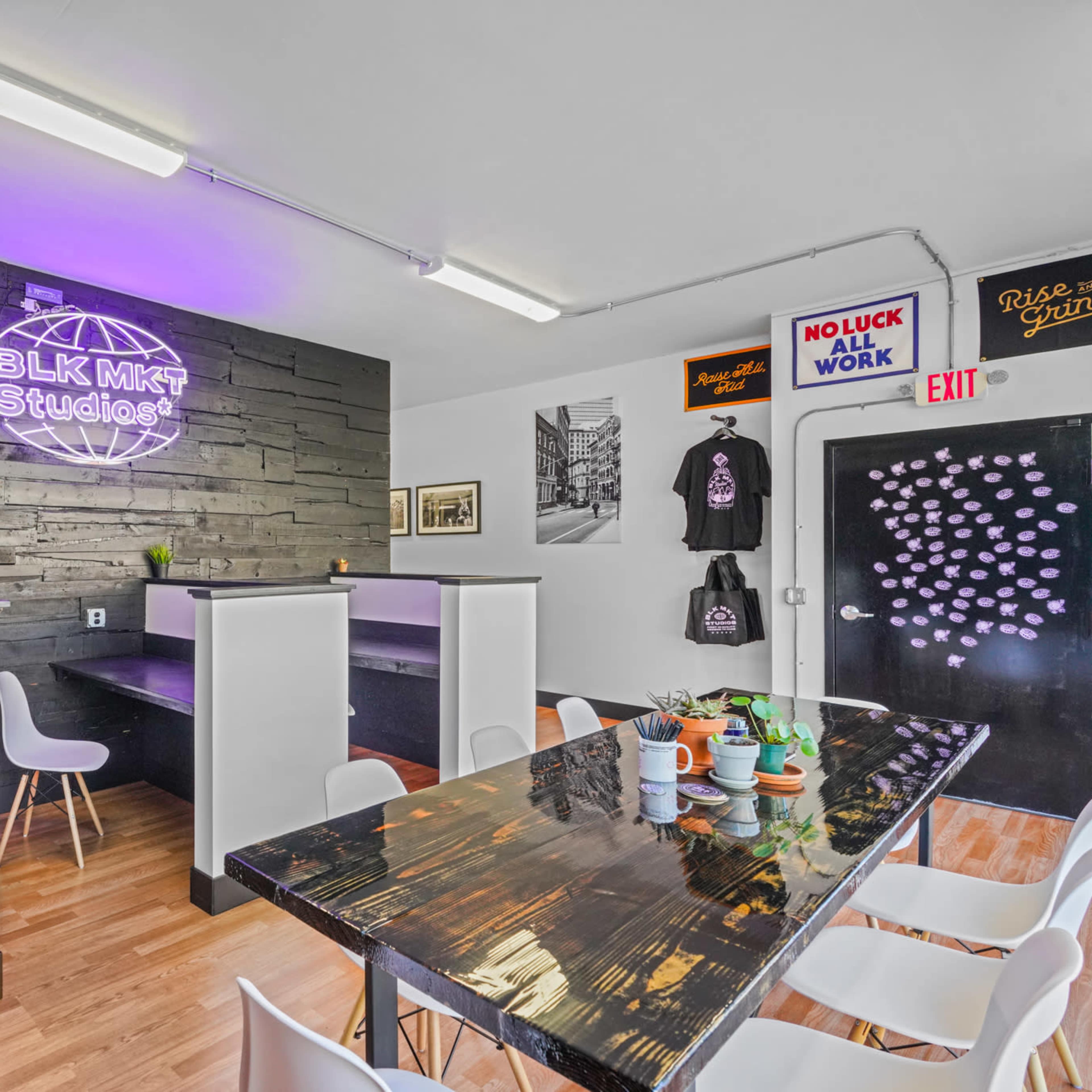 A modern studio space with a black wooden accent wall, a neon sign, a long table set for a meeting, and minimalist white chairs.