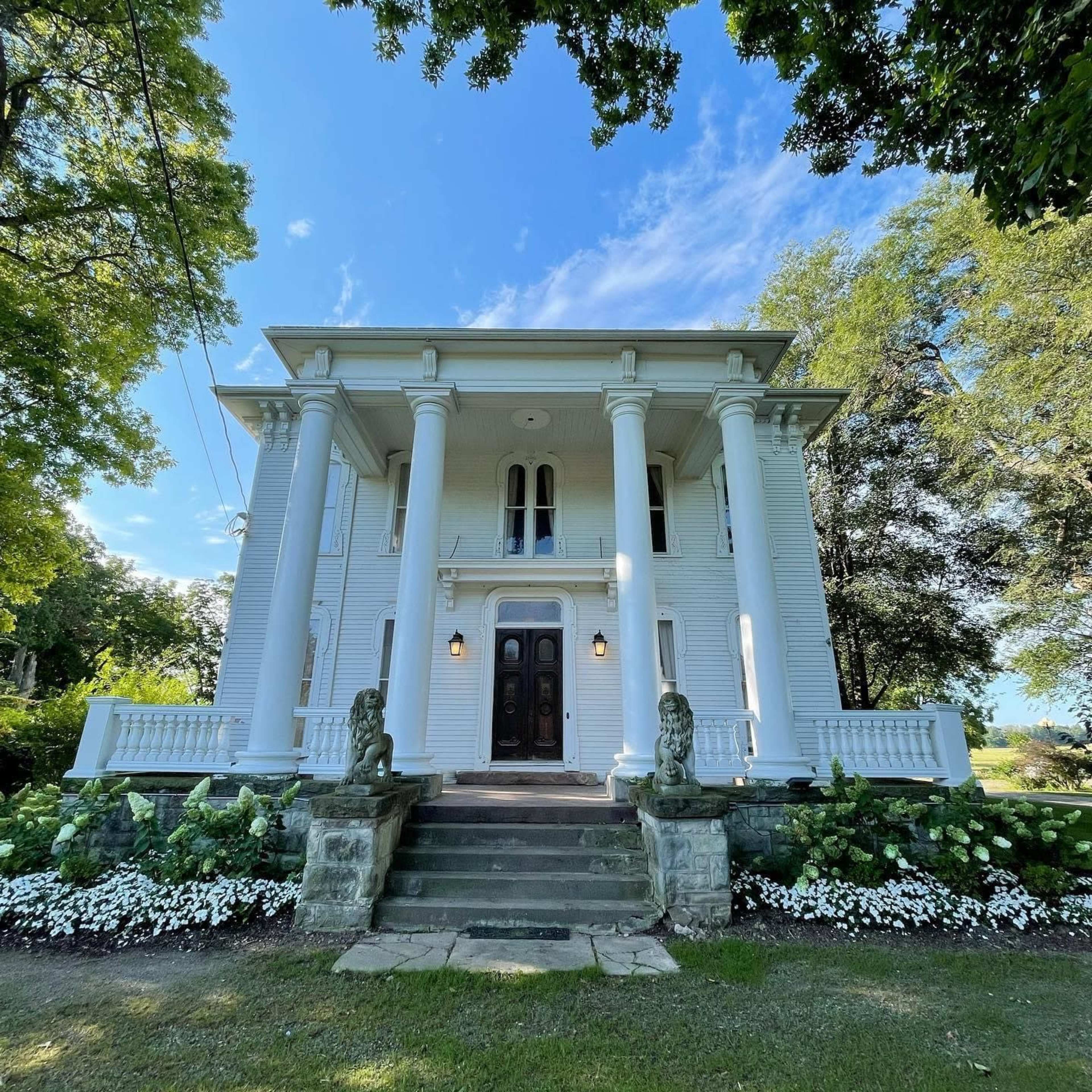 A large white colonial-style house with four prominent columns at the entrance, surrounded by greenery and flowers.