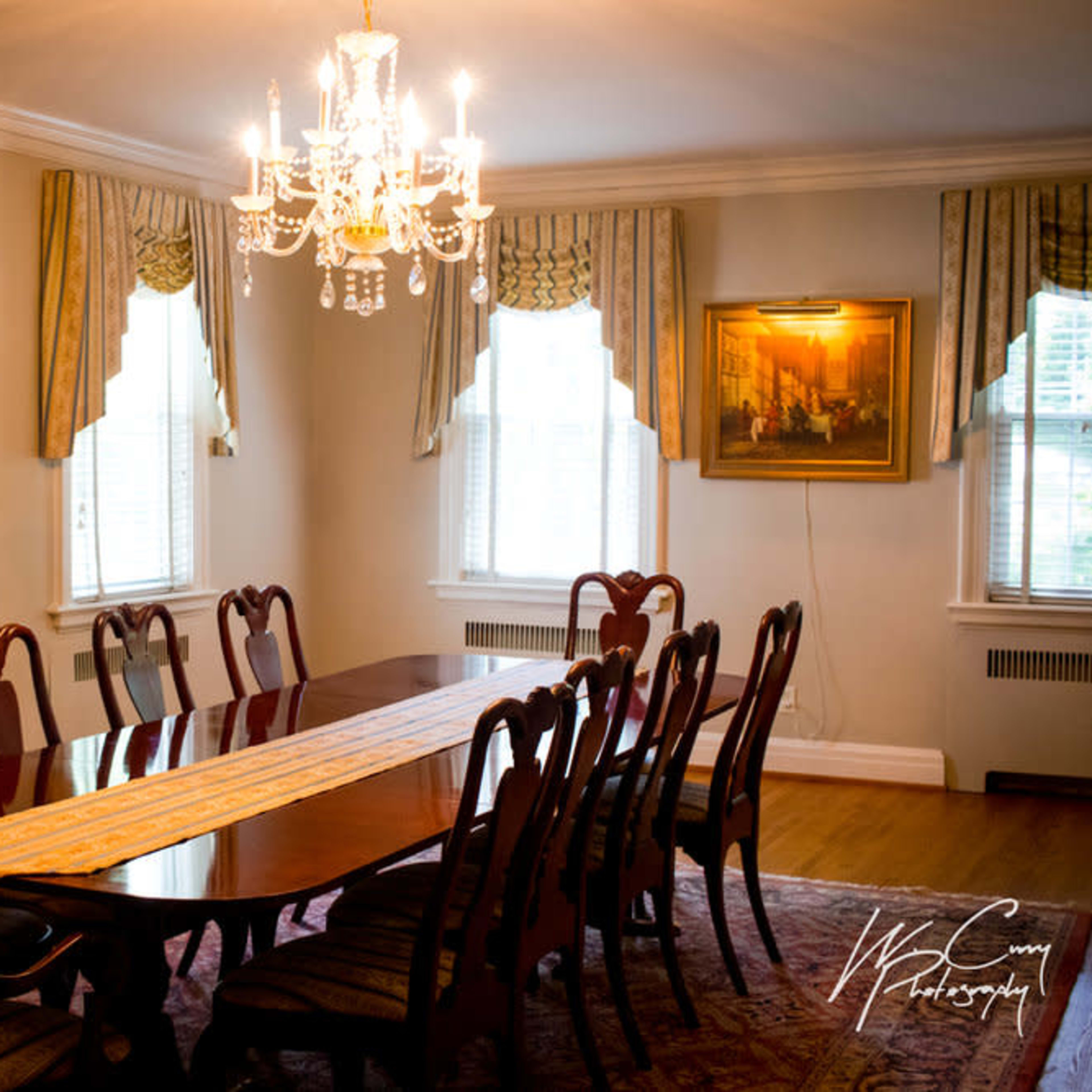 A formal dining room features a long wooden table surrounded by twelve chairs, illuminated by a chandelier and adorned with artwork on the walls.