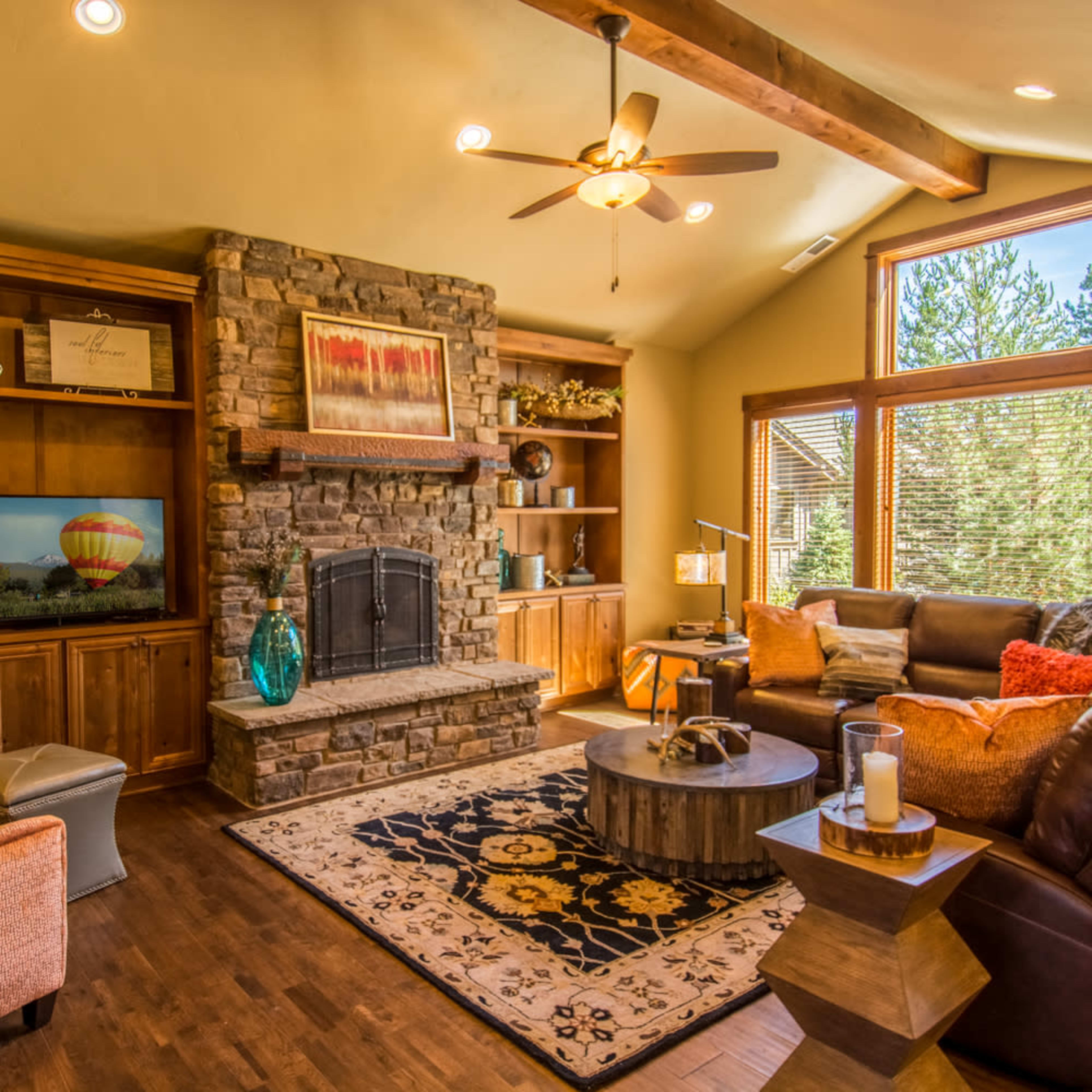 A cozy living room with a stone fireplace, wooden shelves, a large round coffee table, and ample natural light coming through large windows.