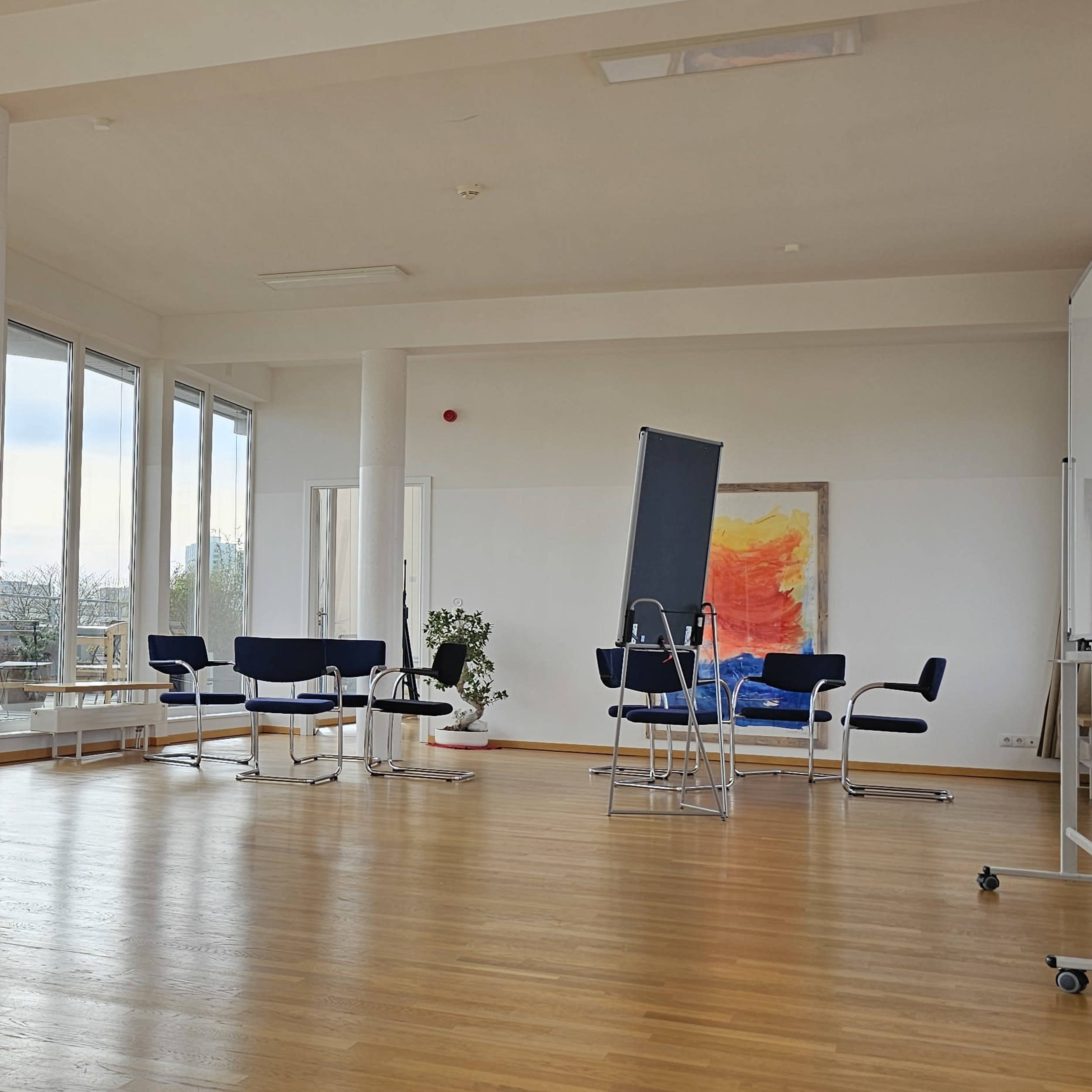 A person stands near large windows in a bright, spacious room with wooden floors and modern furniture.