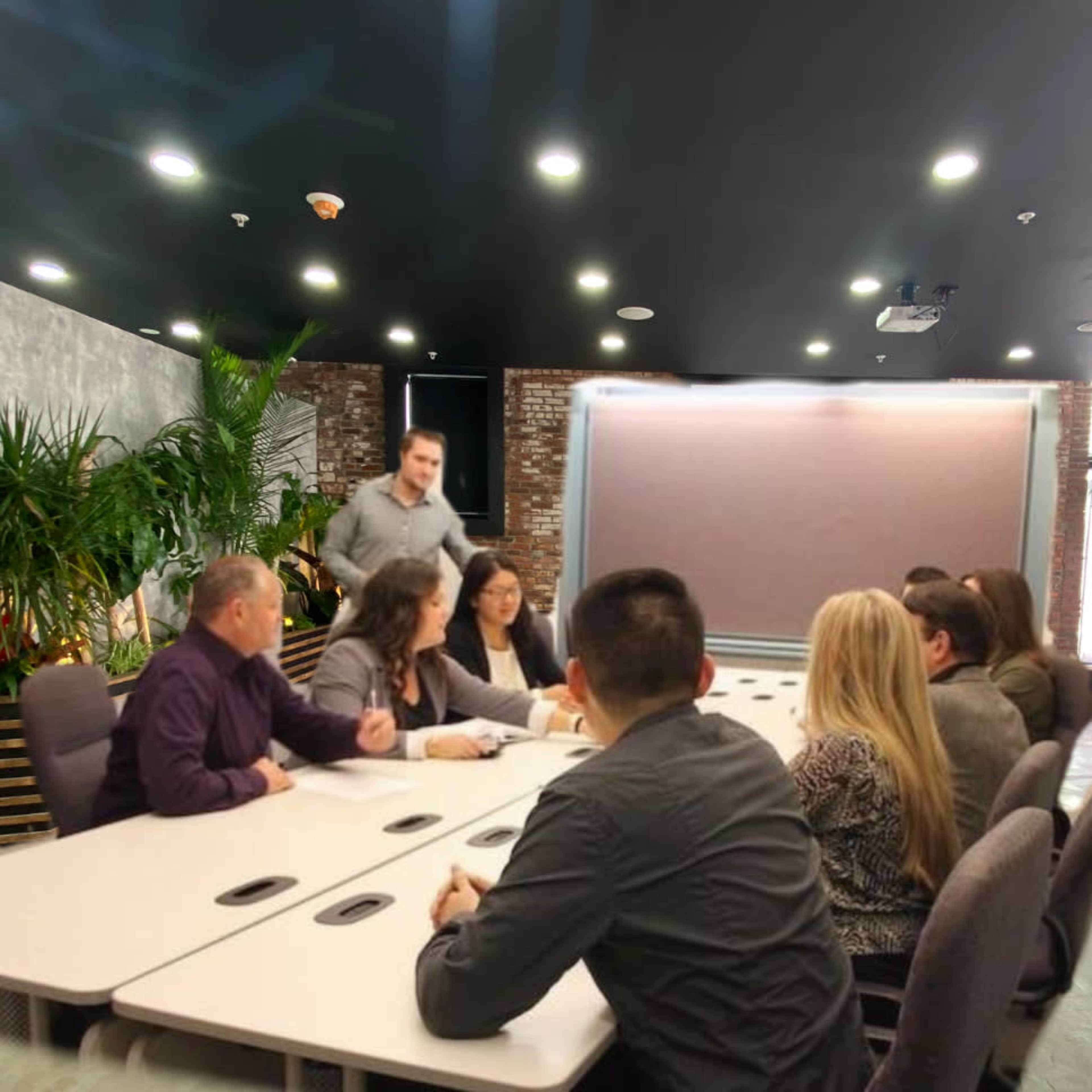 A group of eight people is seated around a long conference table in a well-lit meeting room with plants and a large screen in the background.