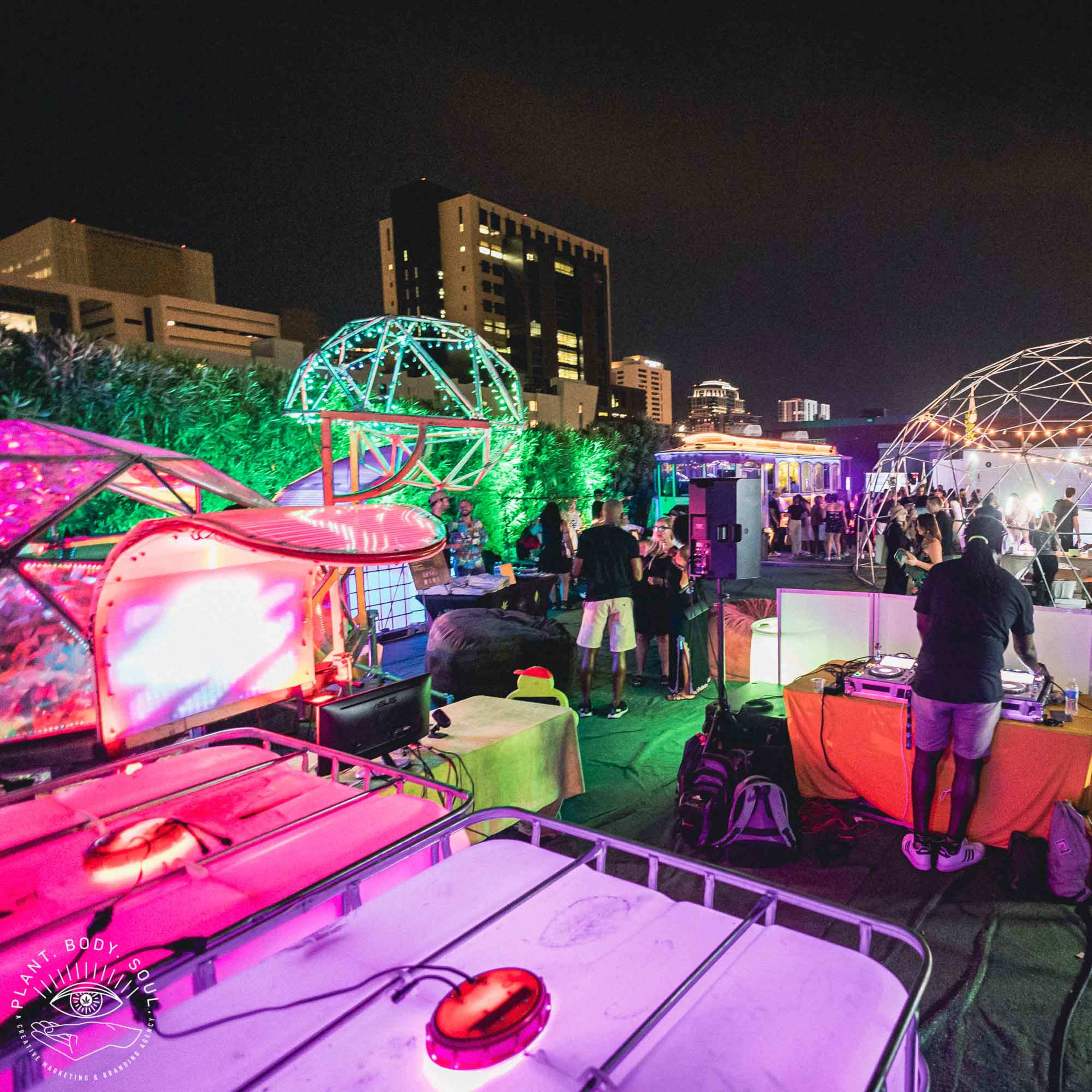 A vibrant outdoor event takes place at night, featuring illuminated geodesic domes, a DJ booth, and attendees mingling on a green carpeted area.