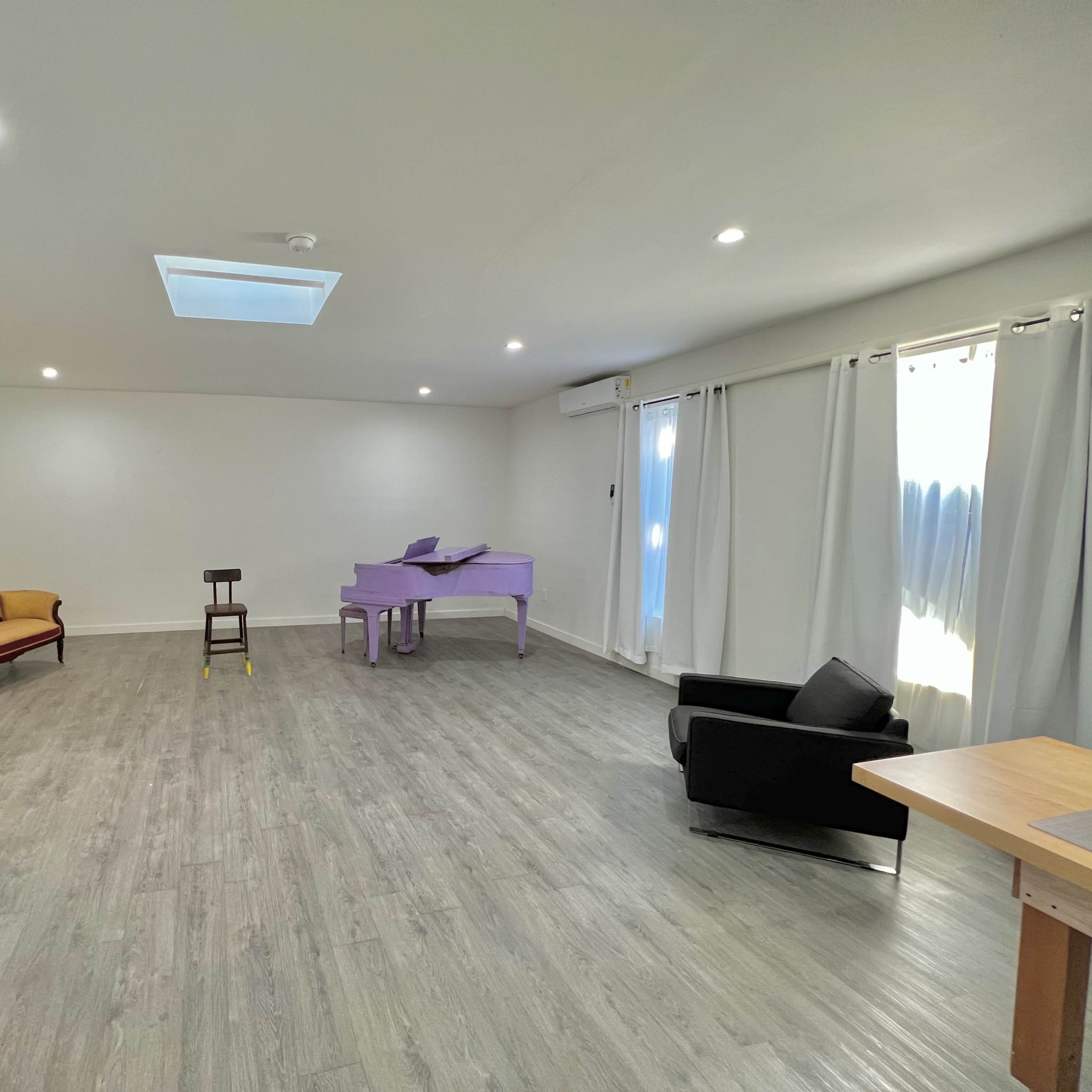 A spacious room with light-colored flooring, featuring a purple piano, a wooden chair, a vintage sofa, a full-length mirror, and large windows with white curtains.