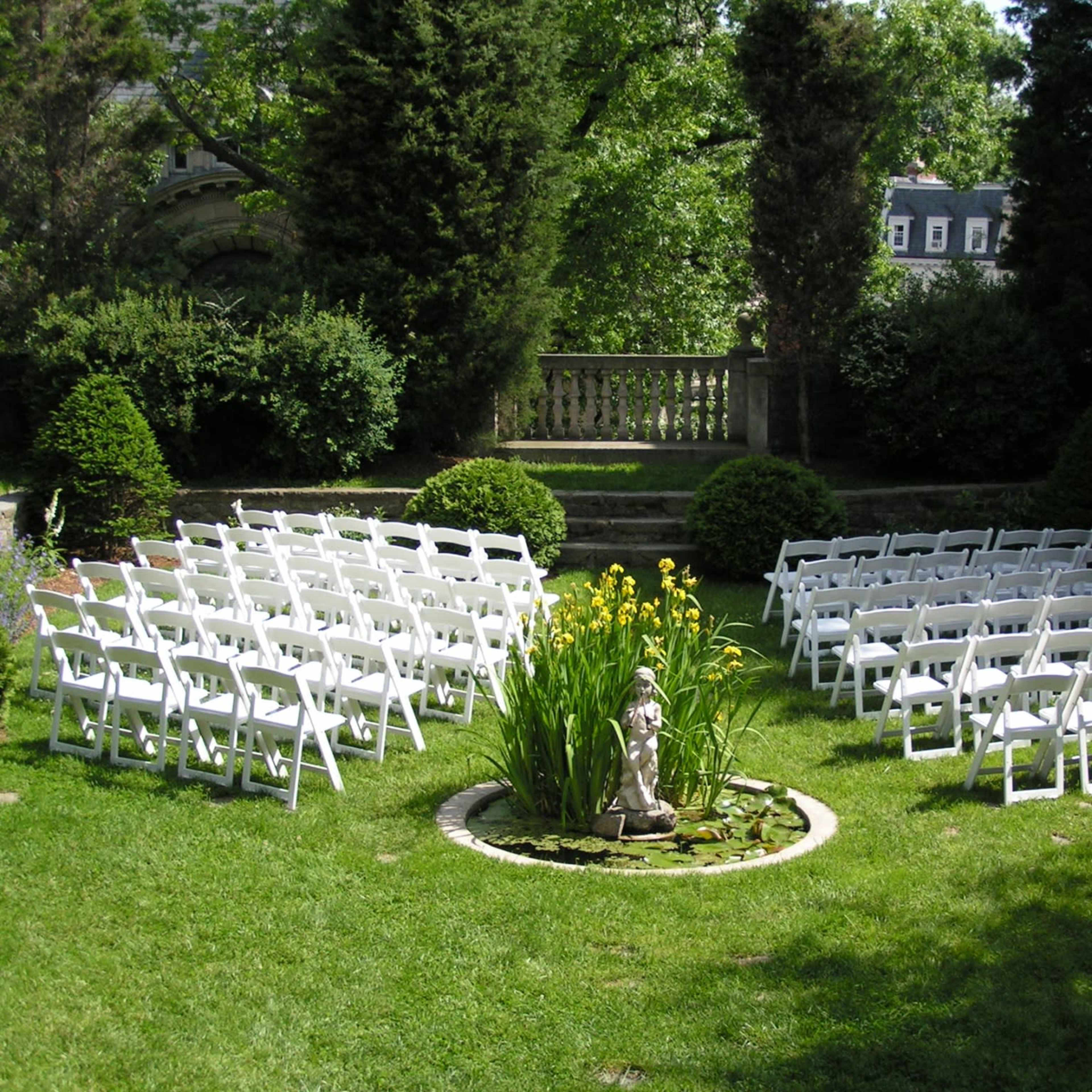 A garden setup with white chairs arranged in rows around a circular flower bed containing a statue and greenery.