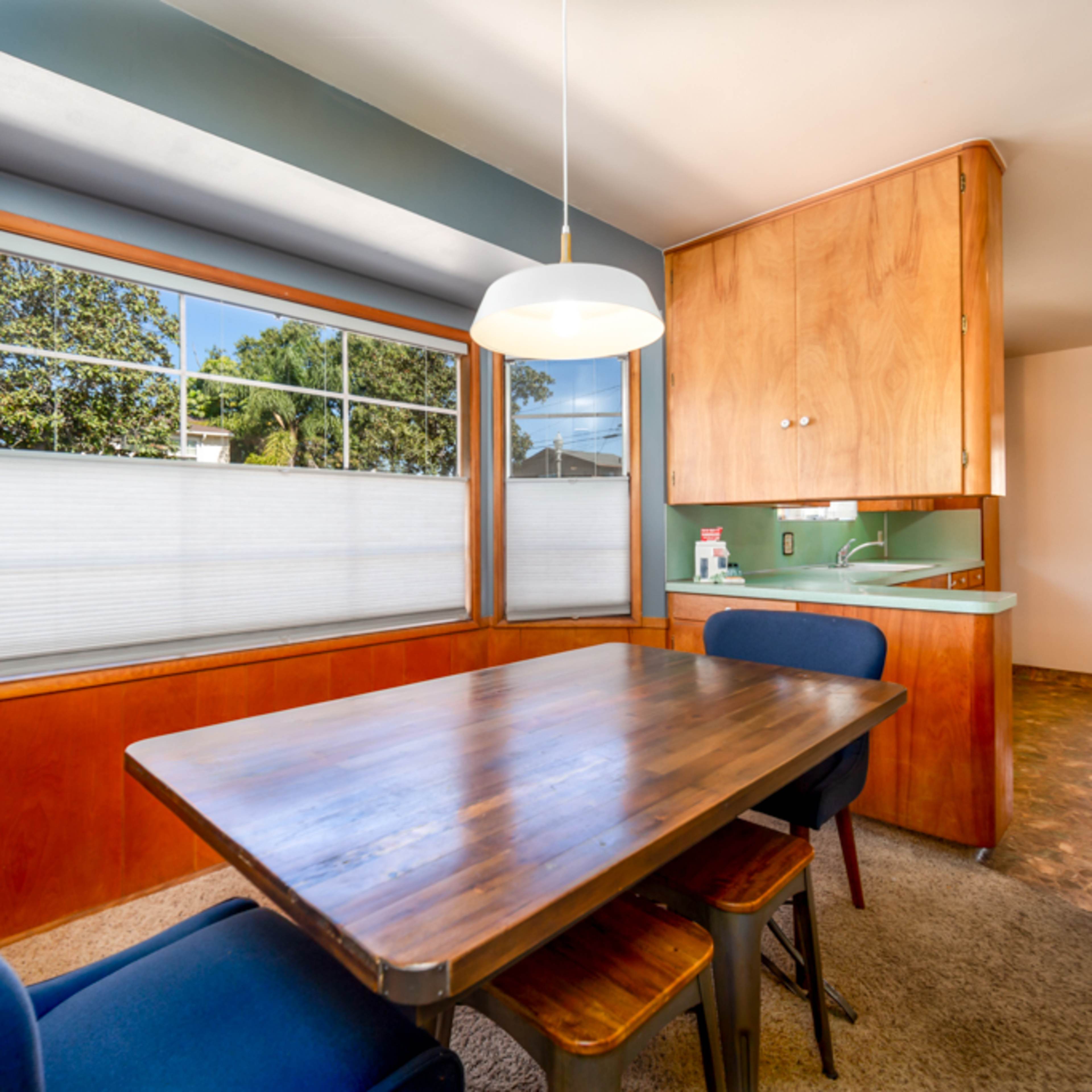 A dining area with a wooden table and blue upholstered chairs, featuring large windows and a kitchen area in the background.