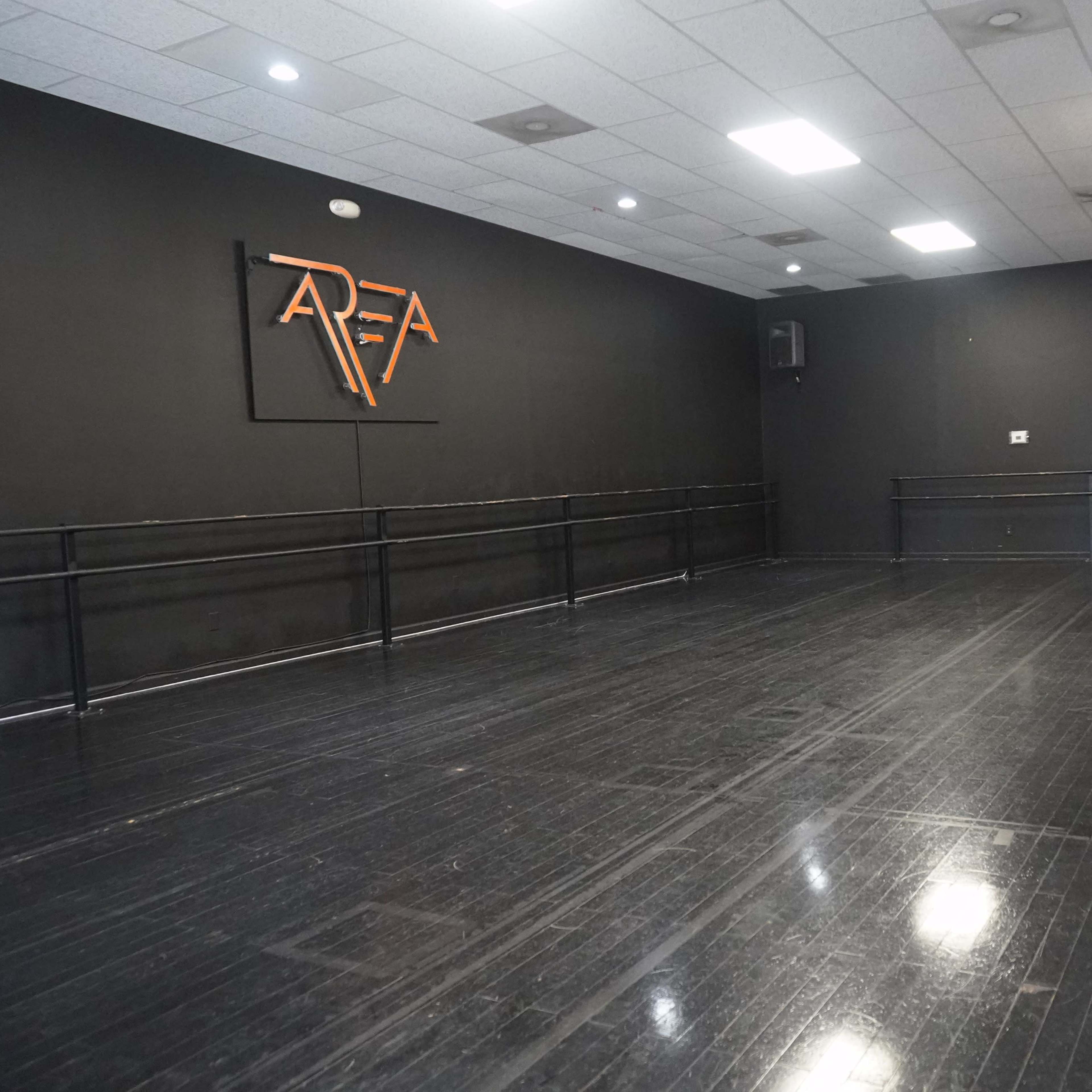 A dance studio featuring a black wall with a logo, wooden flooring, and ballet bars along one side.