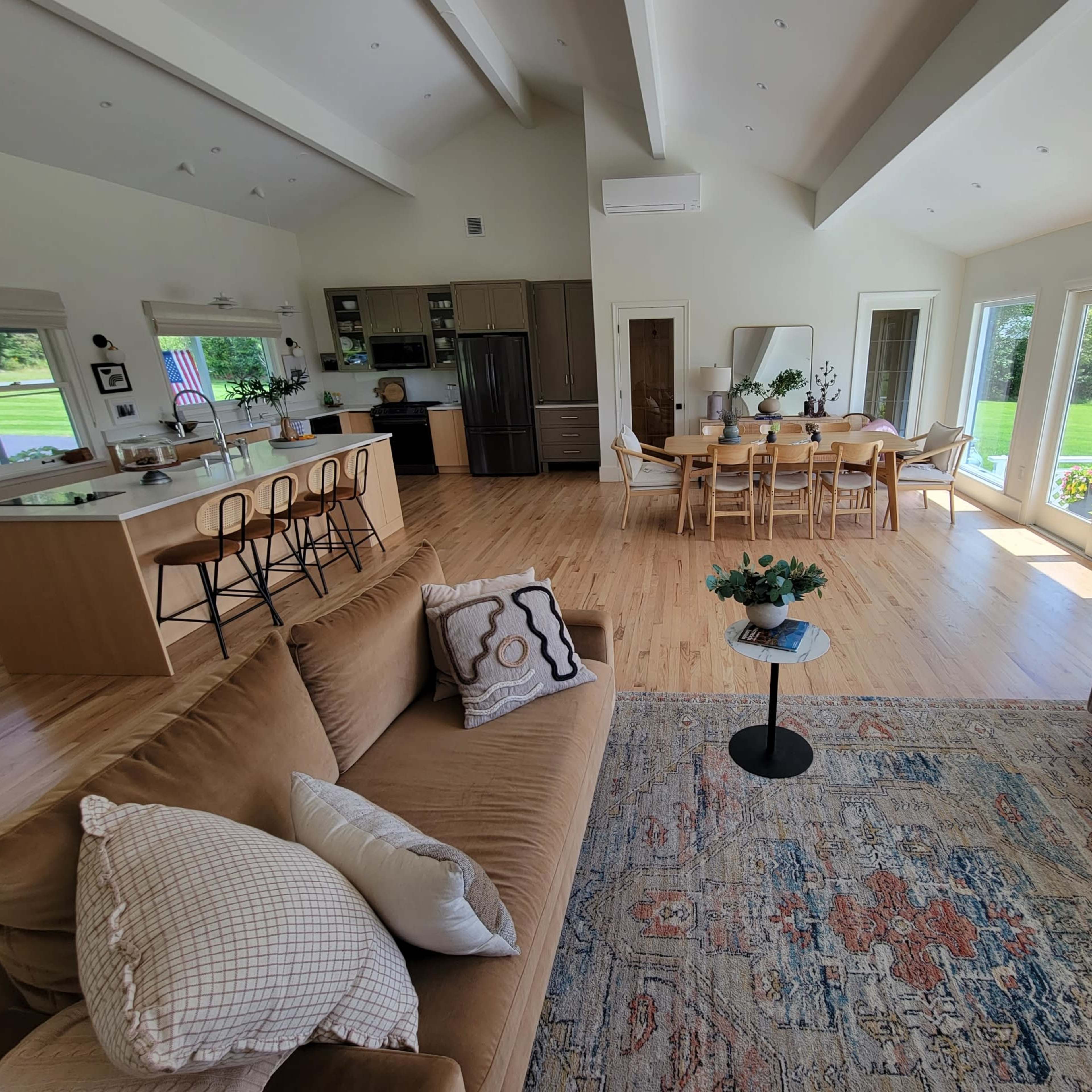 A spacious living area features a sofa, coffee table, and a dining table with chairs, adjoining a kitchen with modern appliances and large windows overlooking a garden.