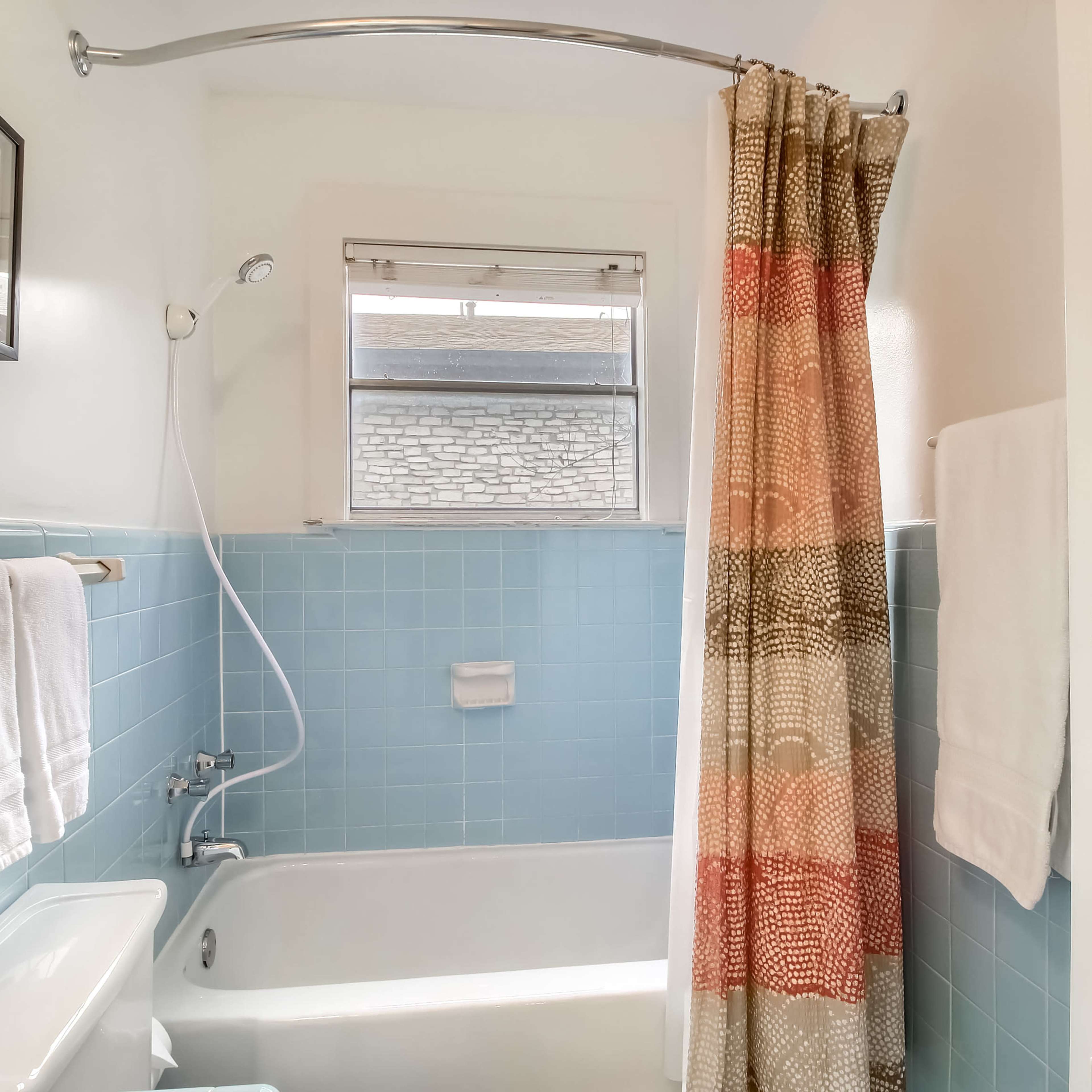 A bathroom with a blue tiled wall, a white bathtub, a shower curtain in orange and beige patterns, and white towels hanging on a rack.
