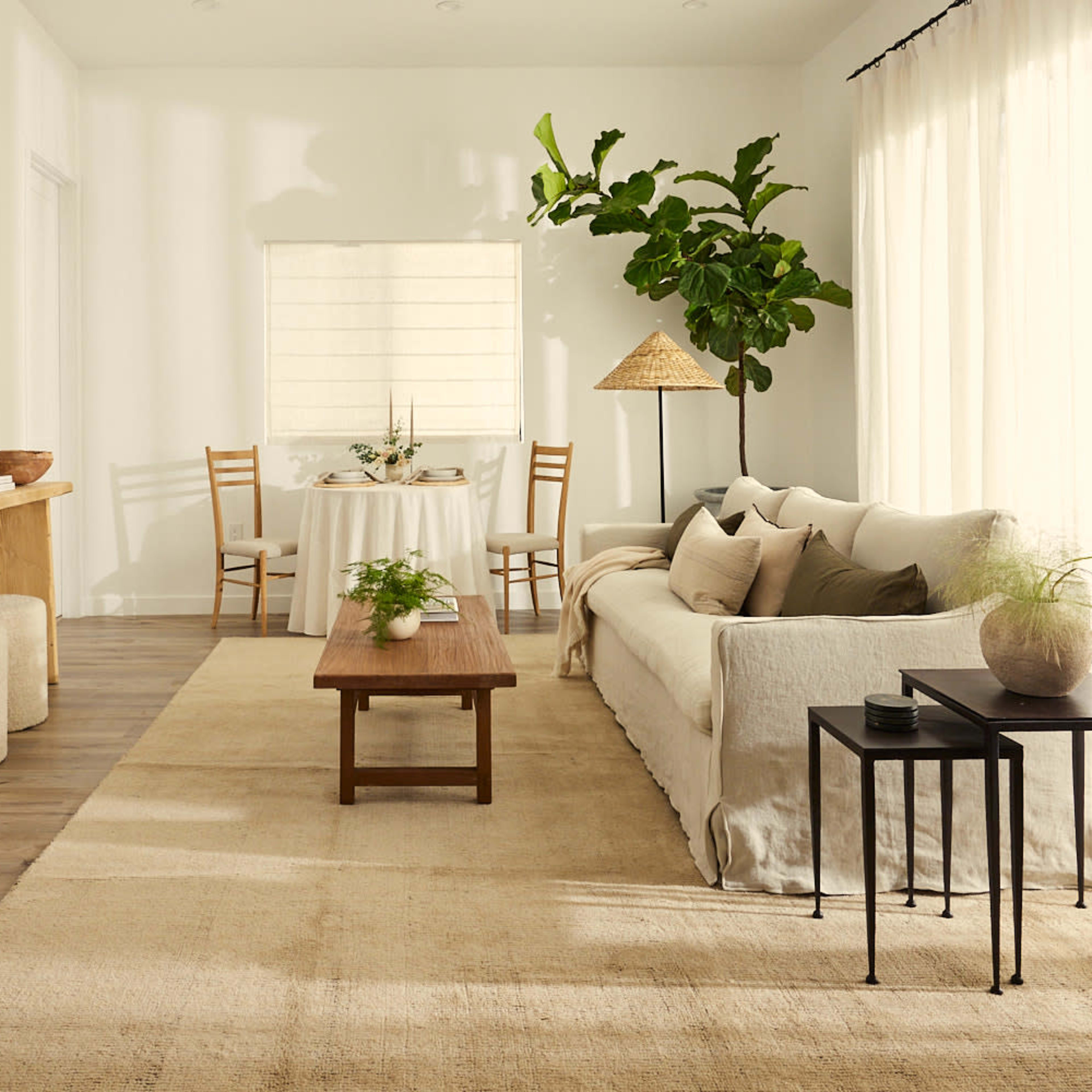 A light-filled living room features a neutral color palette with a sofa, coffee table, two side tables, and a dining area in the background.
