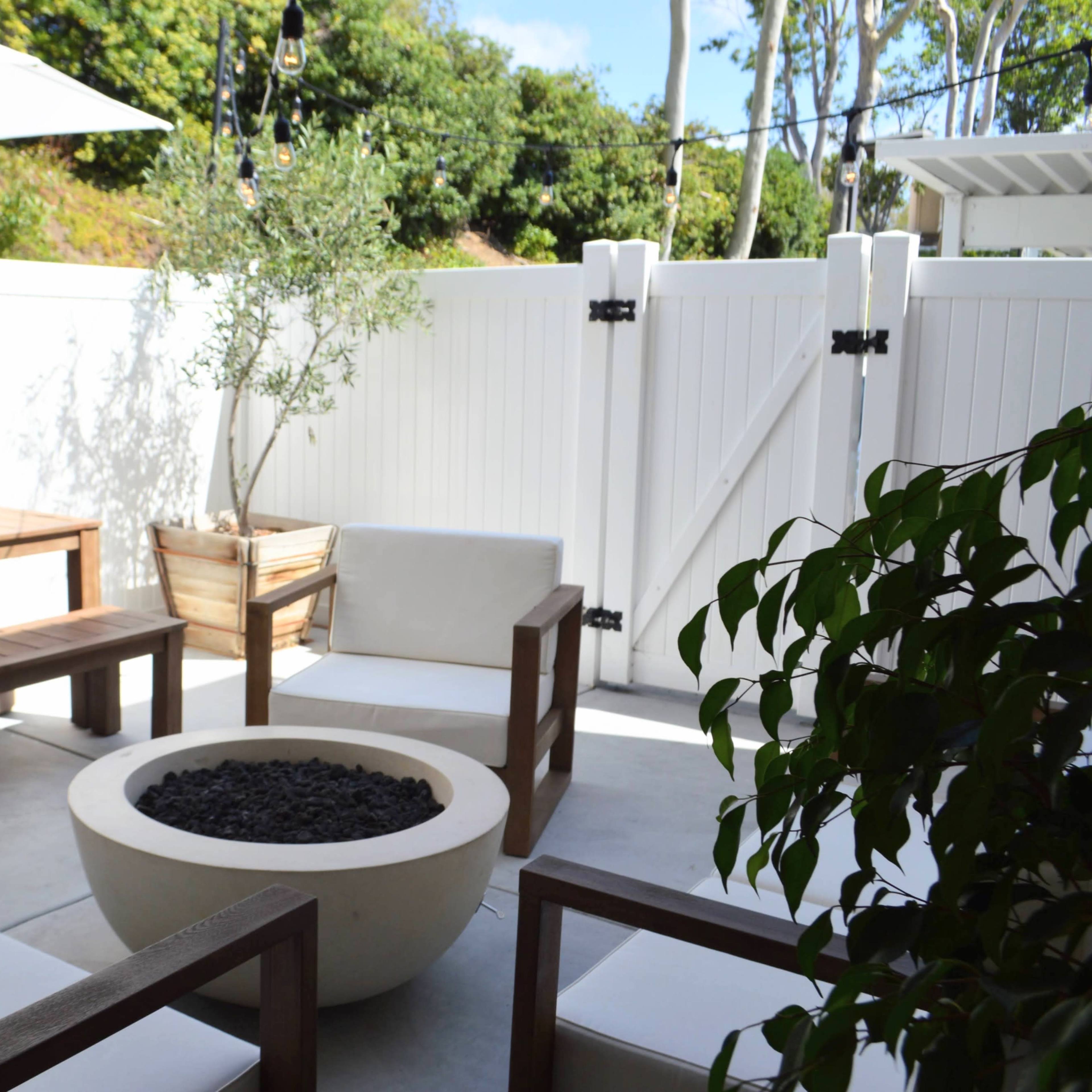 A patio area features a wooden table, four chairs with white cushions, and a circular fire pit surrounded by a fenced garden.