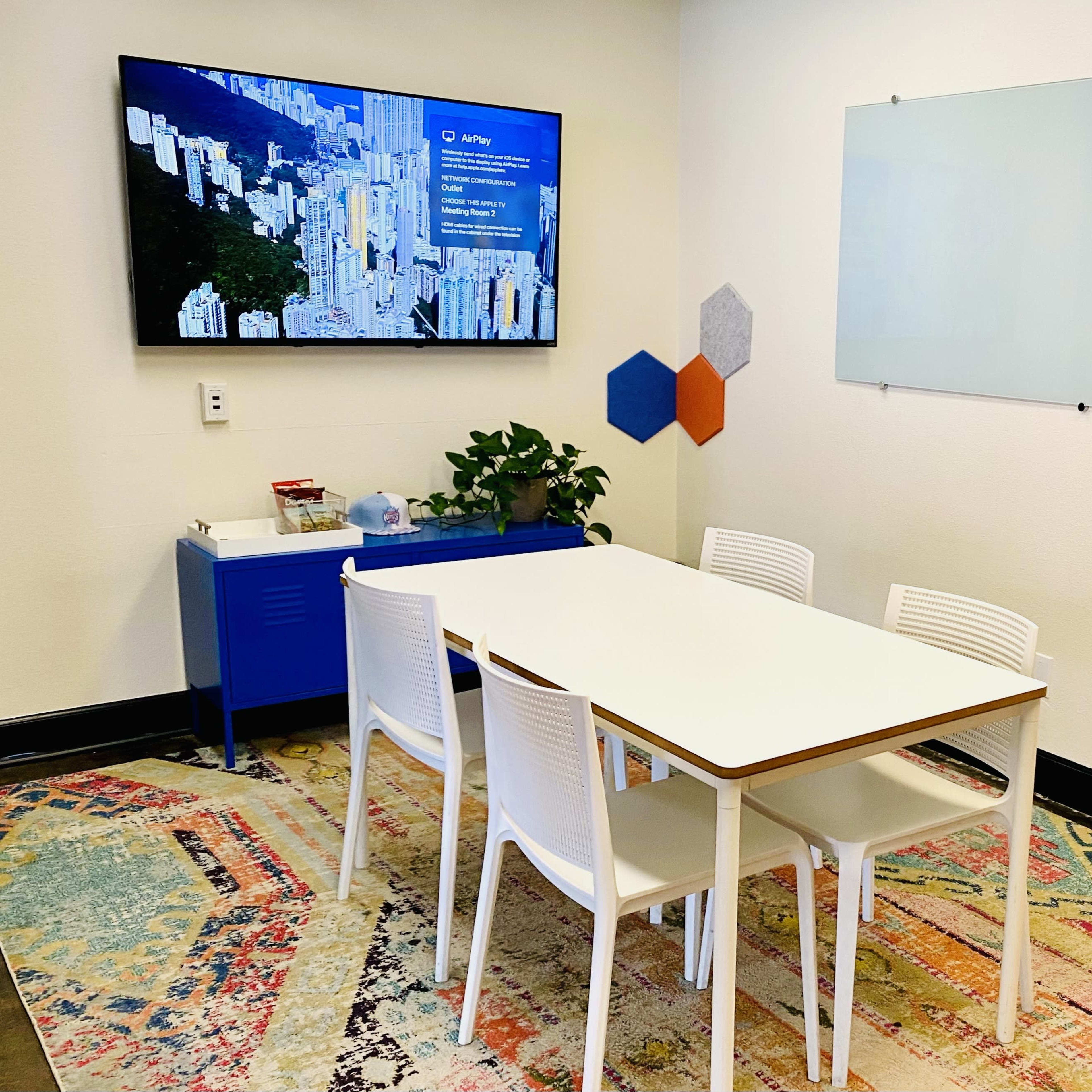 A conference room features a white table surrounded by four white chairs, a blue cabinet, and a television displaying a cityscape, with two empty whiteboards on the wall.
