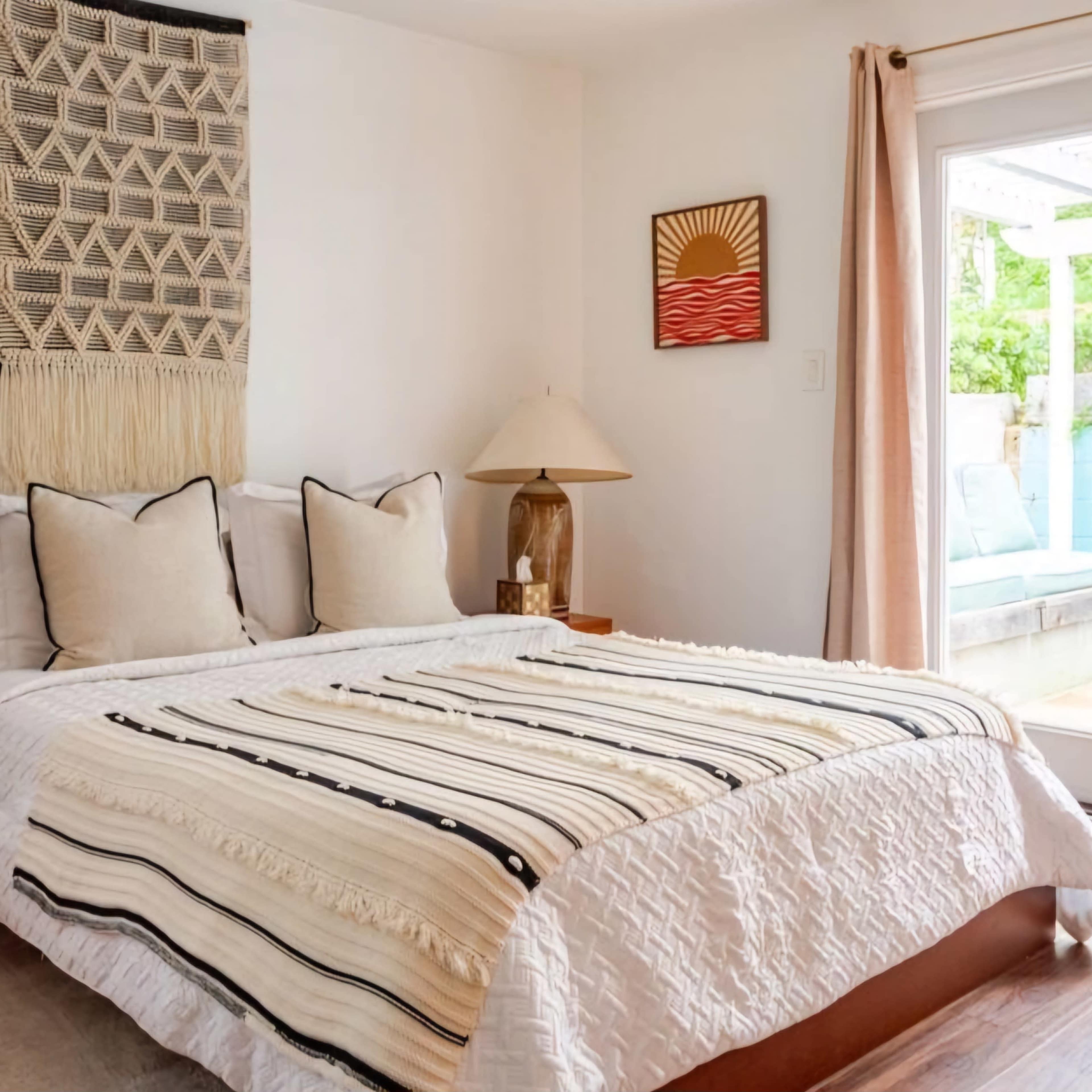 A cozy bedroom with a bed featuring a patterned blanket, decorative pillows, and a wall hanging above the headboard, along with a light-filled balcony accessible through sliding glass doors.