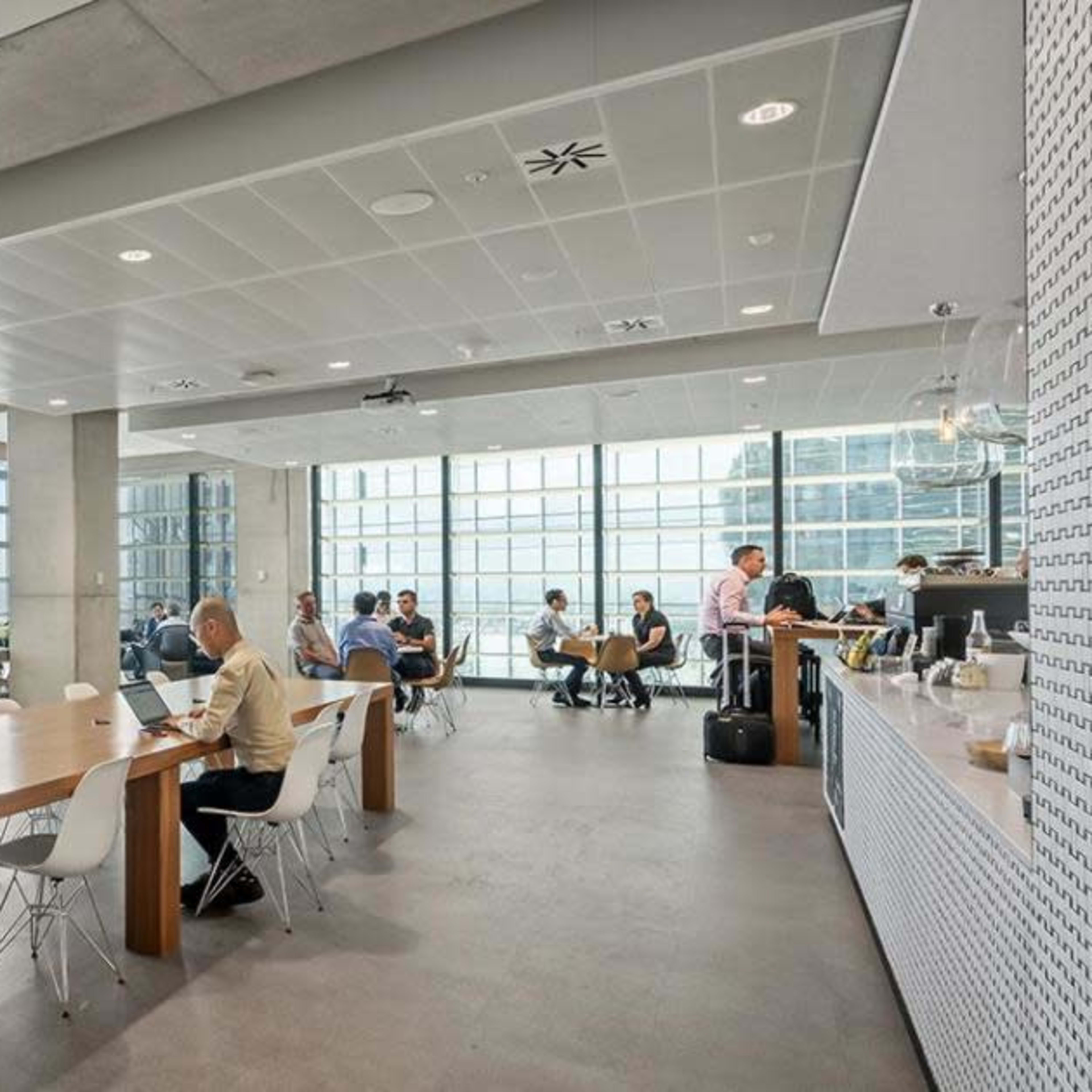 A modern office space features a long table with chairs, large windows showcasing a city view, and several workers engaged in various activities.