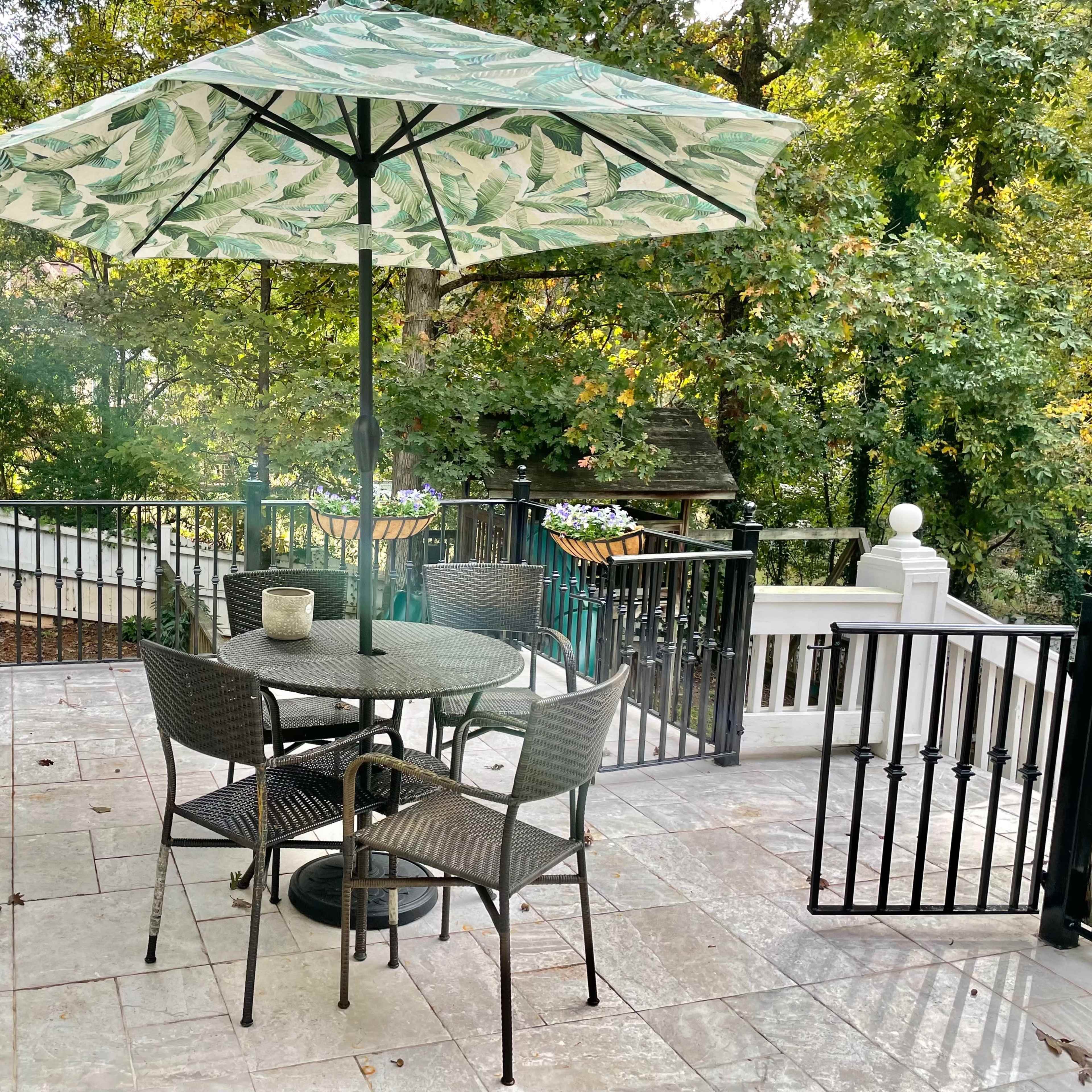 A patio area features a round table with four chairs beneath a green patterned umbrella, surrounded by trees and a railing.