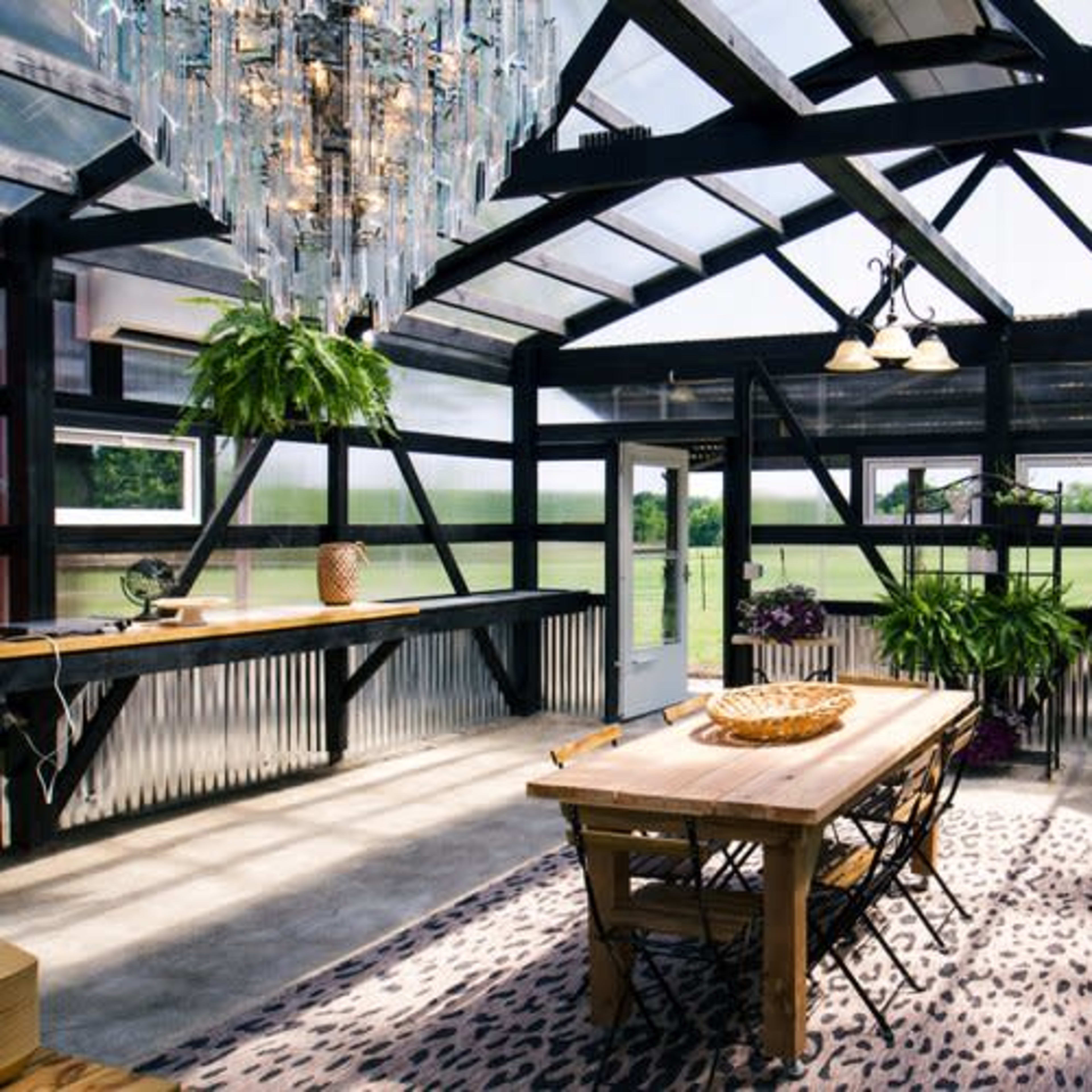 A spacious greenhouse with a wooden dining table, surrounded by plants and natural light streaming through large windows.