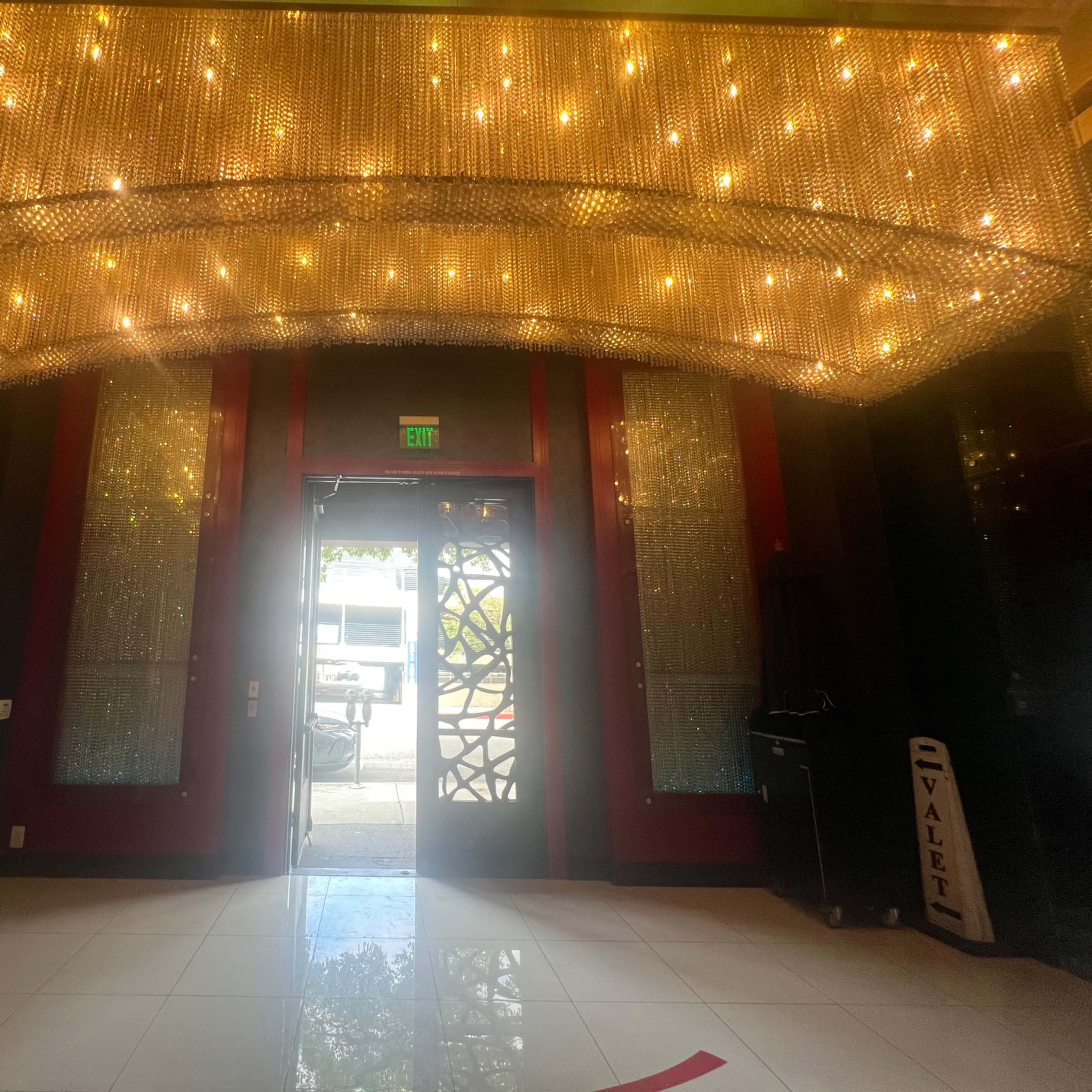 A brightly lit entrance area featuring a large chandelier, glass doors, and a valet station to the side.