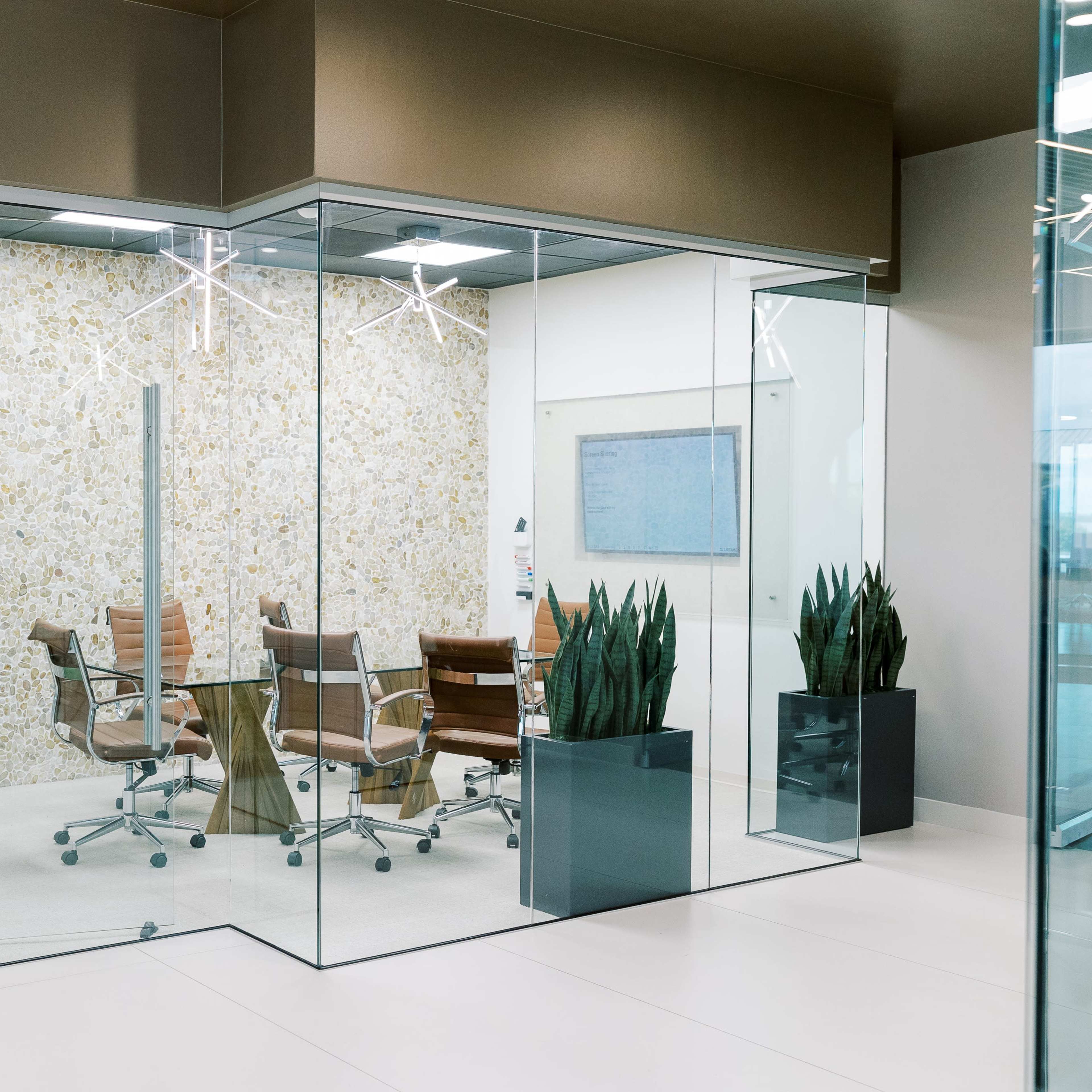 A glass-walled conference room features a wooden table surrounded by ergonomic chairs and is decorated with indoor plants.