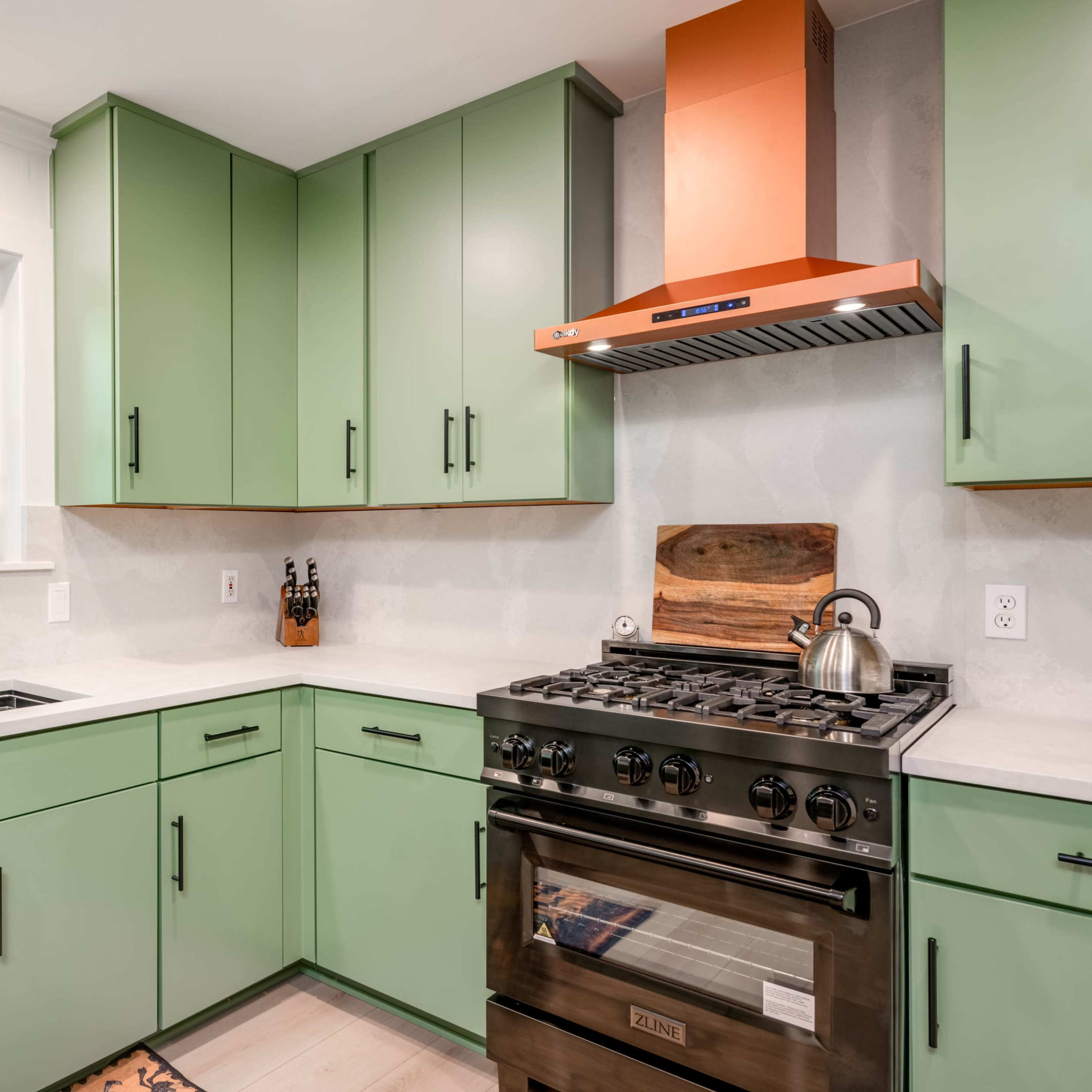 A modern kitchen with green cabinets, a stainless steel gas stove, and a black faucet, complemented by an orange range hood.