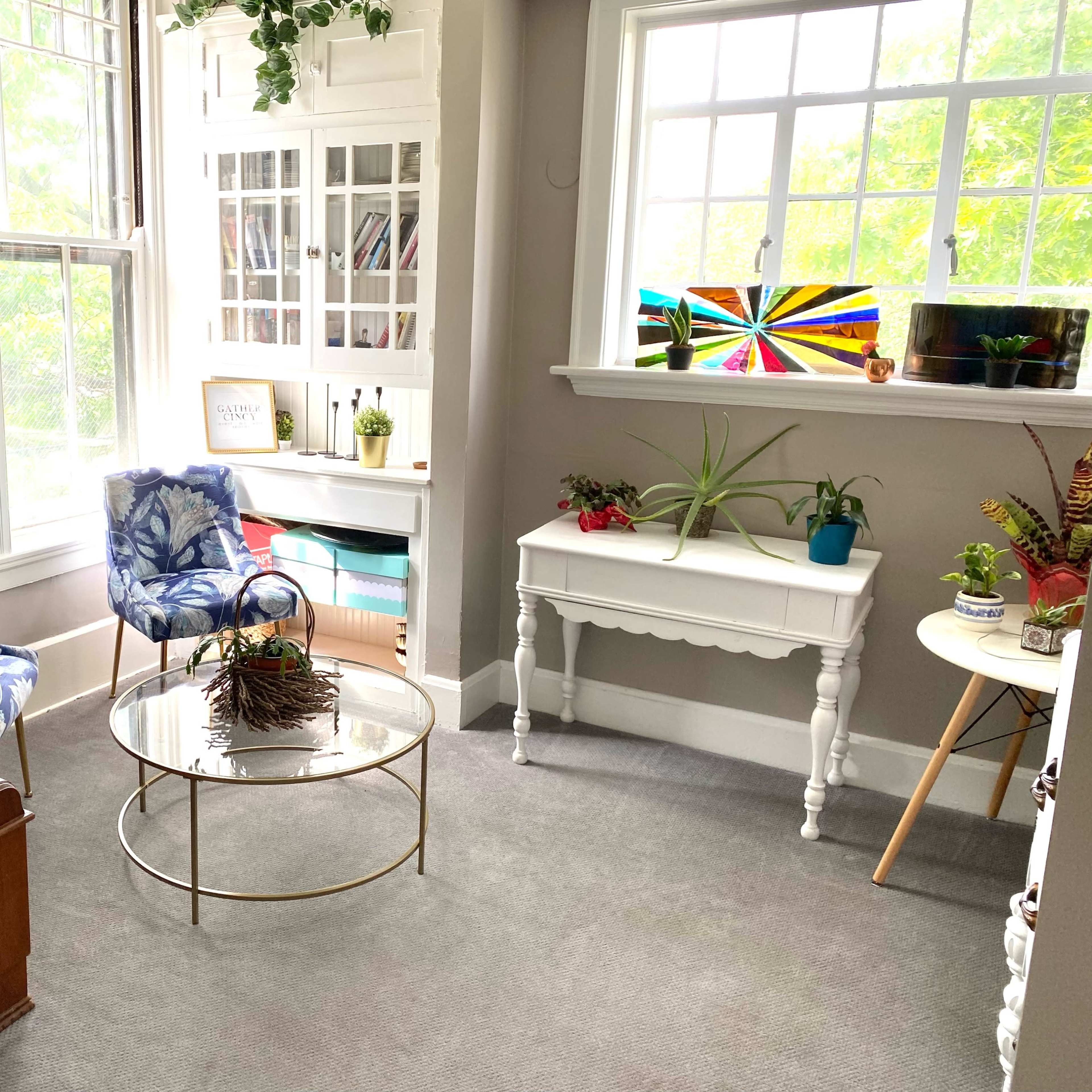 A cozy reading nook features two blue patterned chairs, a glass coffee table, and a white console table with plants, surrounded by large windows.