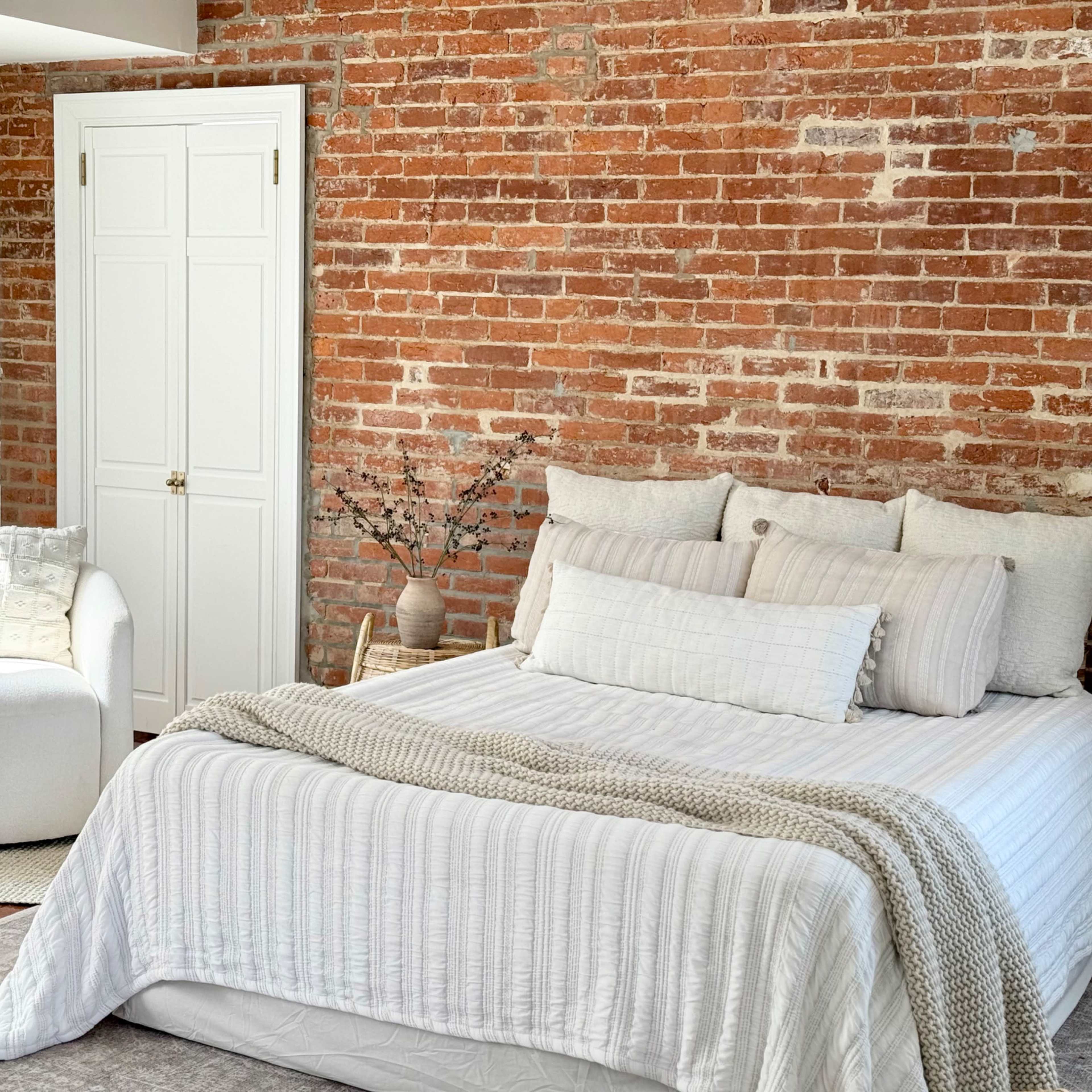 A cozy bedroom features a white bed with layered bedding and a round armchair, set against a brick accent wall.