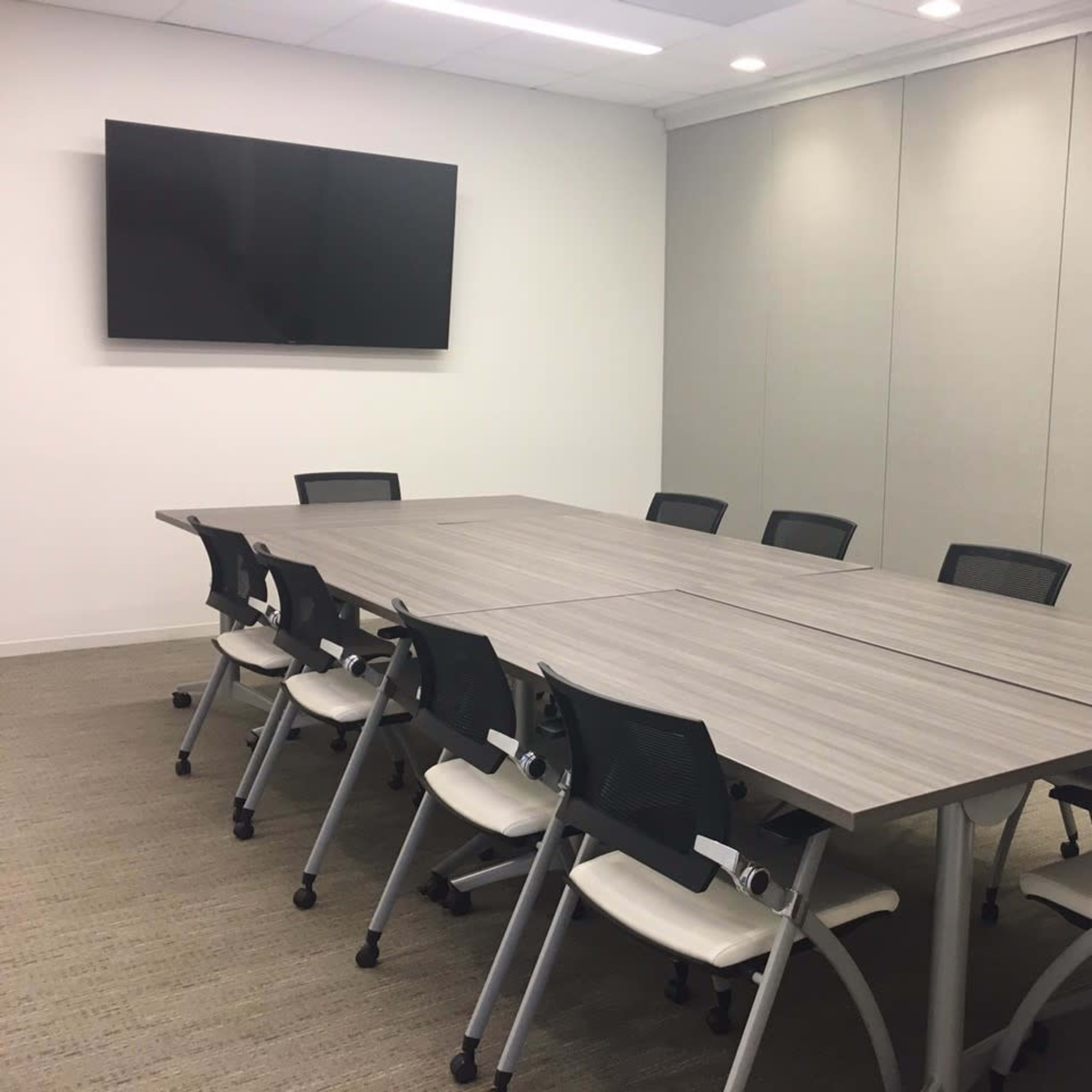 A spacious conference room features a large, rectangular table surrounded by eight chairs, with a wall-mounted television in the background.