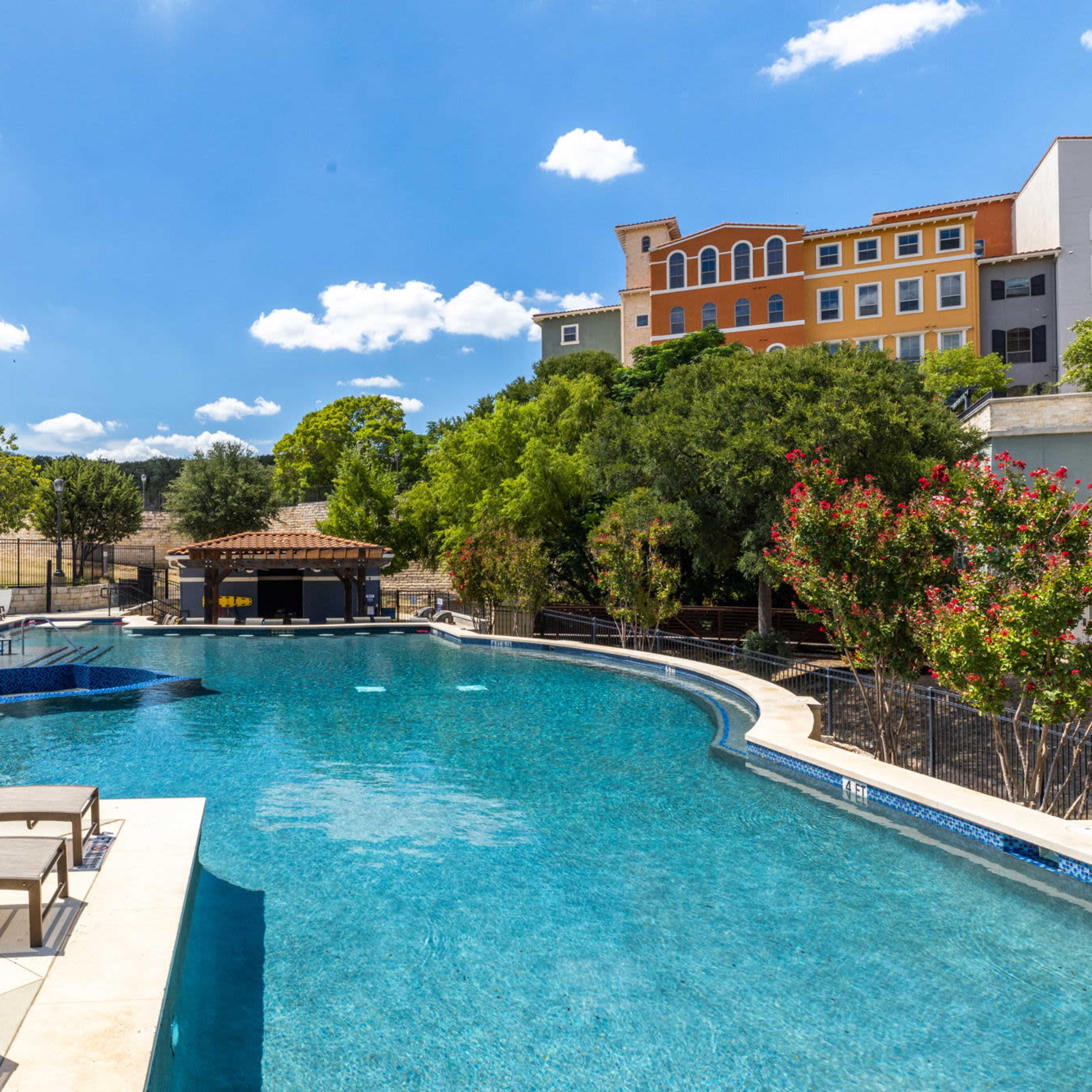 A large swimming pool surrounded by lounge chairs and trees, with a multi-story building in the background under a clear blue sky.