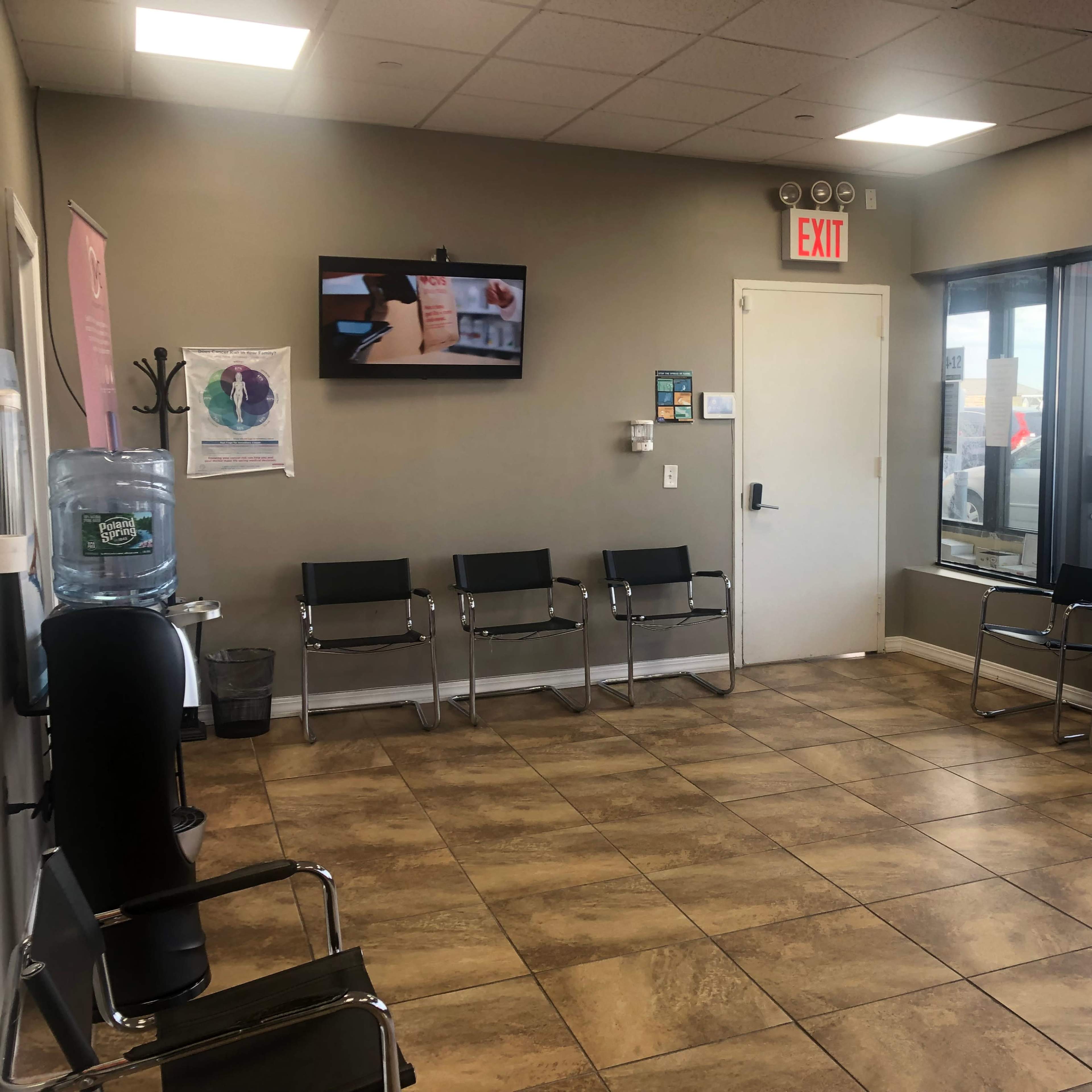 A waiting room with a row of metal chairs, a water cooler, a television on the wall, and large windows providing a view outside.