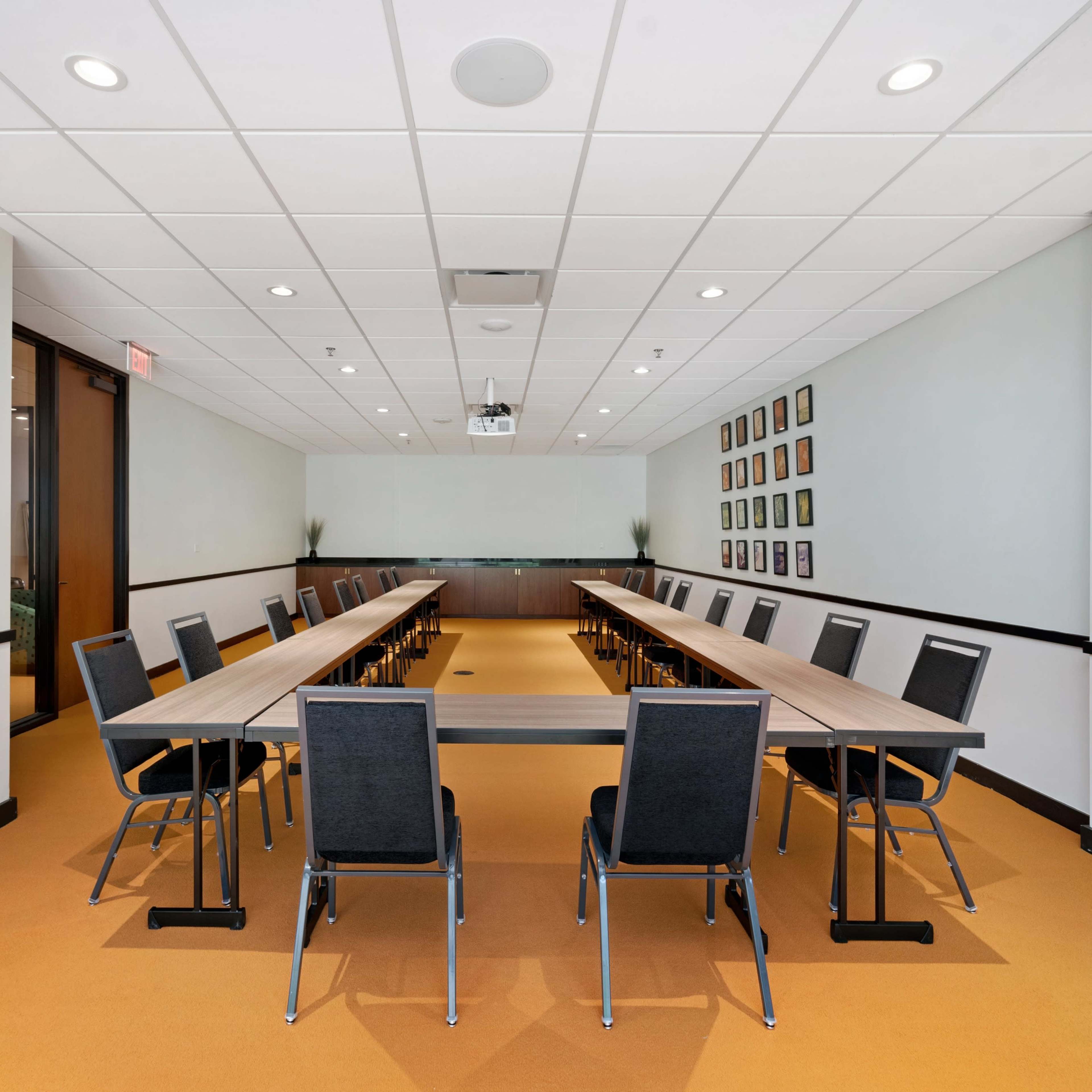 A spacious meeting room features a long table surrounded by chairs, with artwork displayed on the walls and a projector mounted on the ceiling.