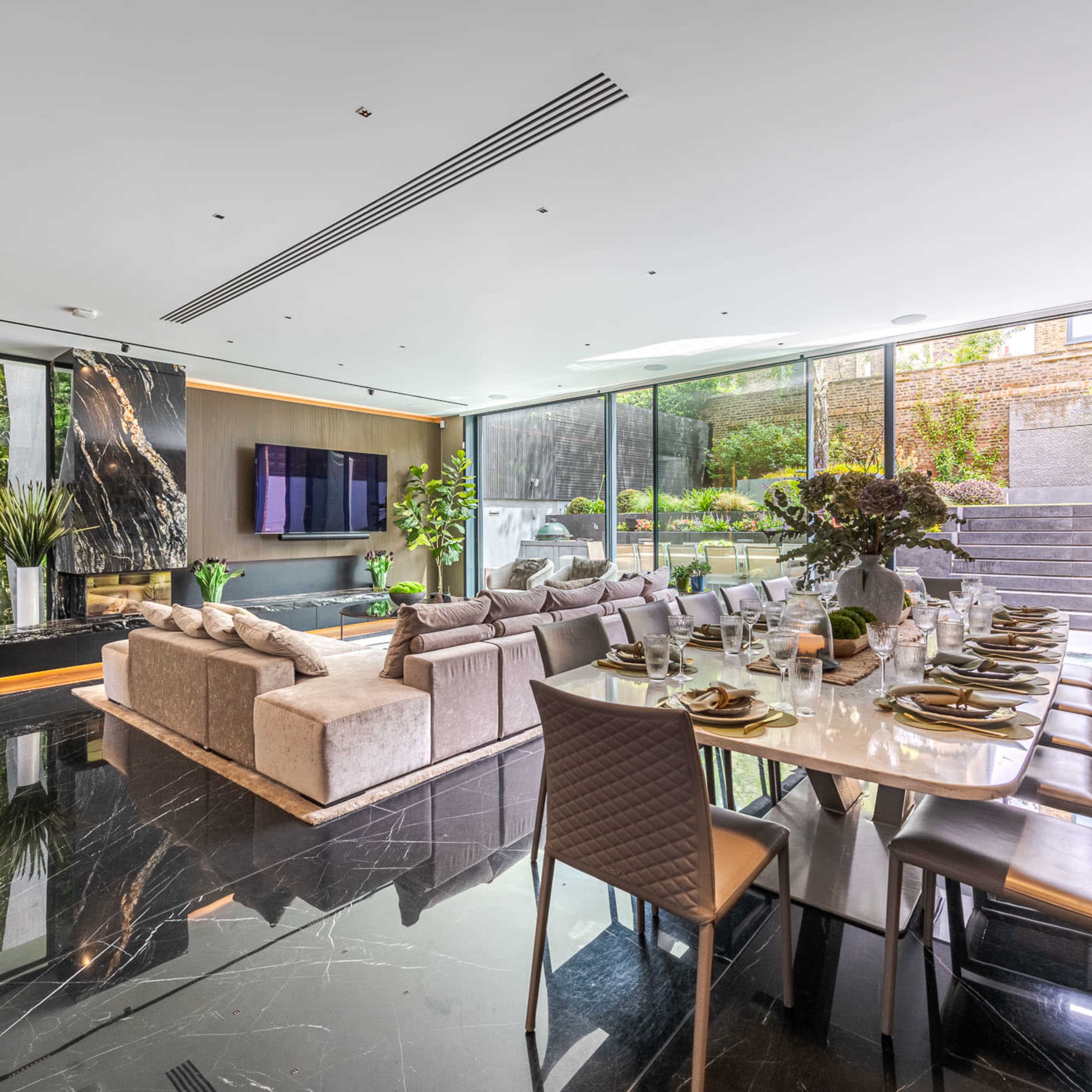 A modern living and dining area features sleek furniture, dark marble flooring, and large windows overlooking a garden.