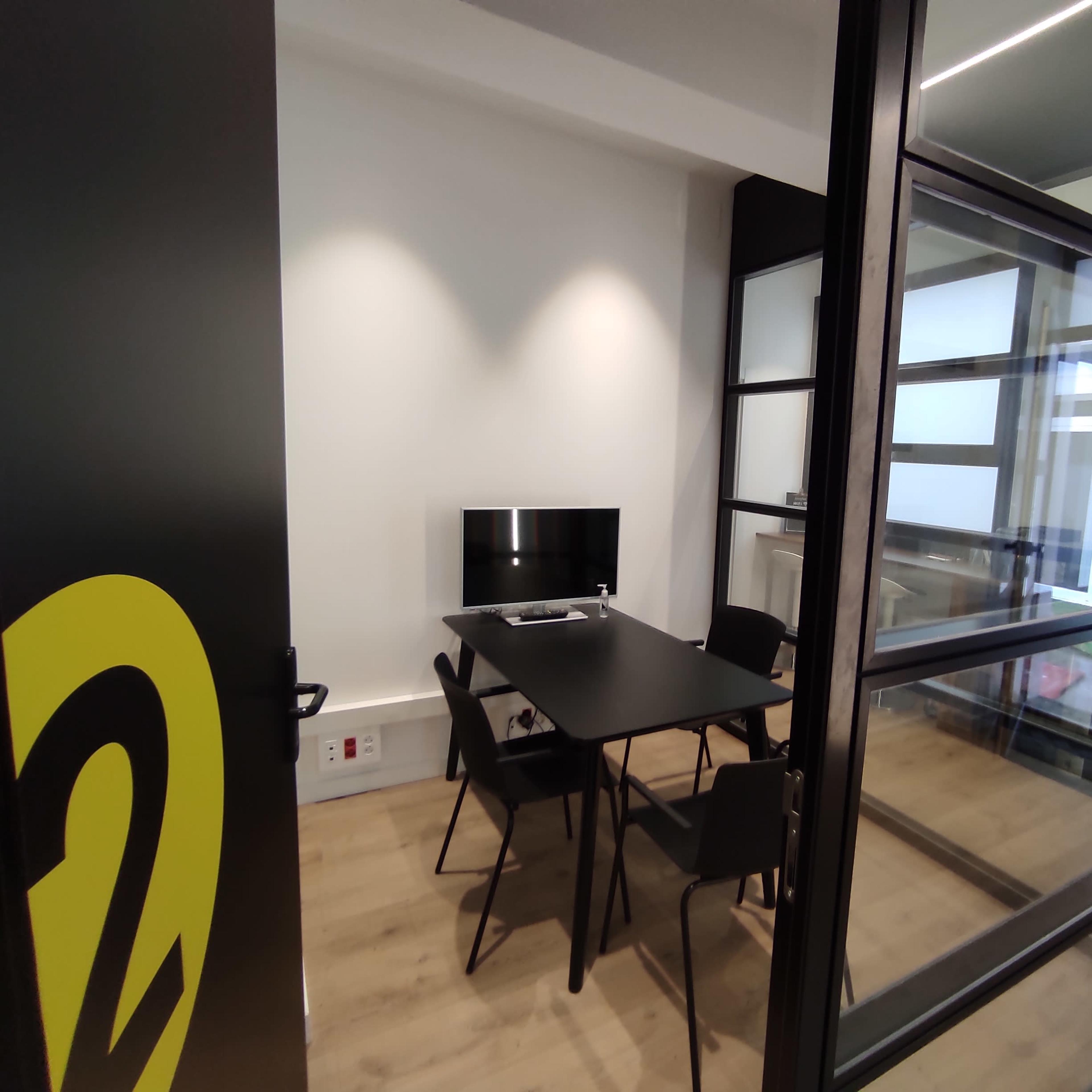 A modern meeting room with a black table and chairs, a small monitor on the table, and large glass panels allowing natural light.