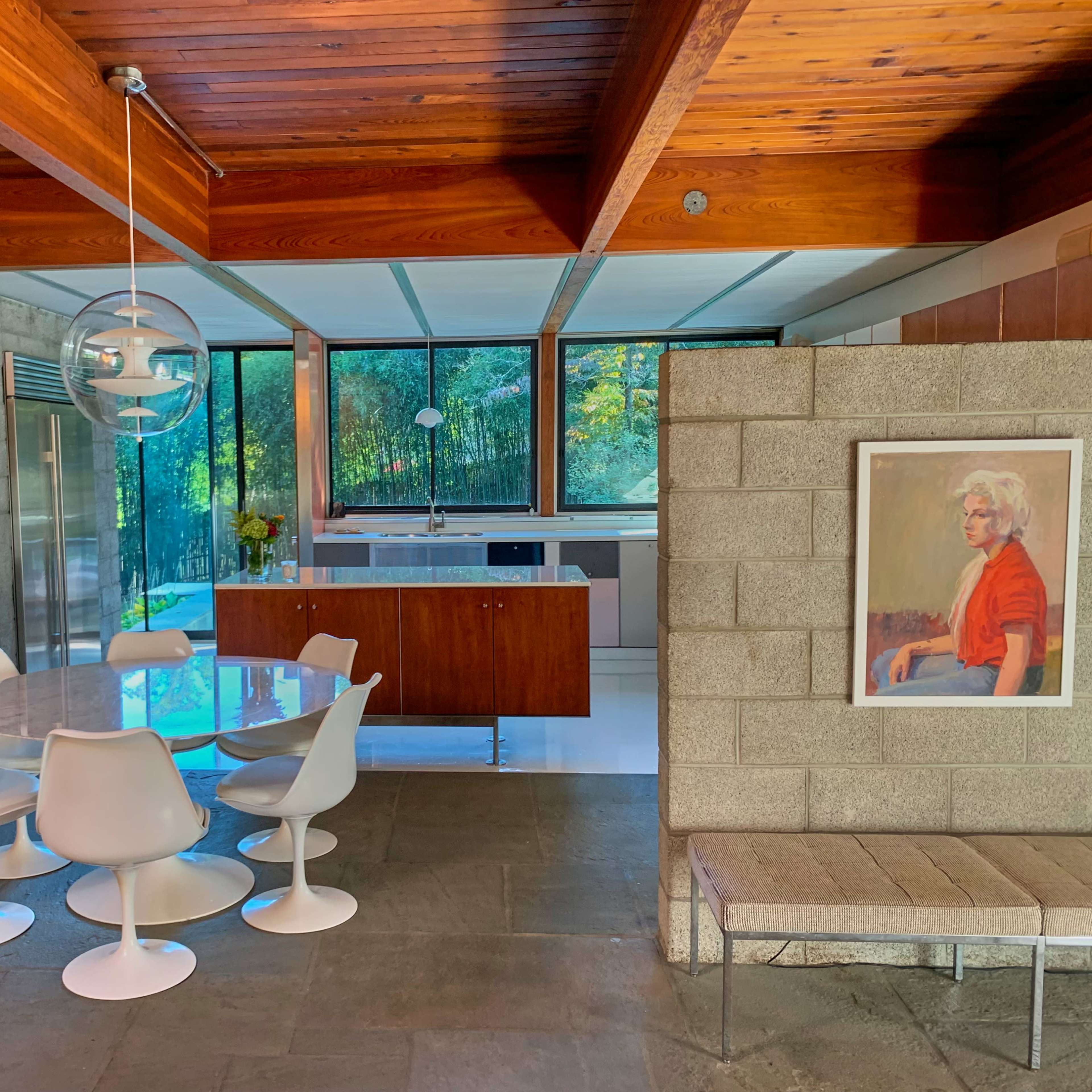 A modern interior features a round dining table surrounded by white chairs, concrete walls, and large windows showcasing a view of greenery.