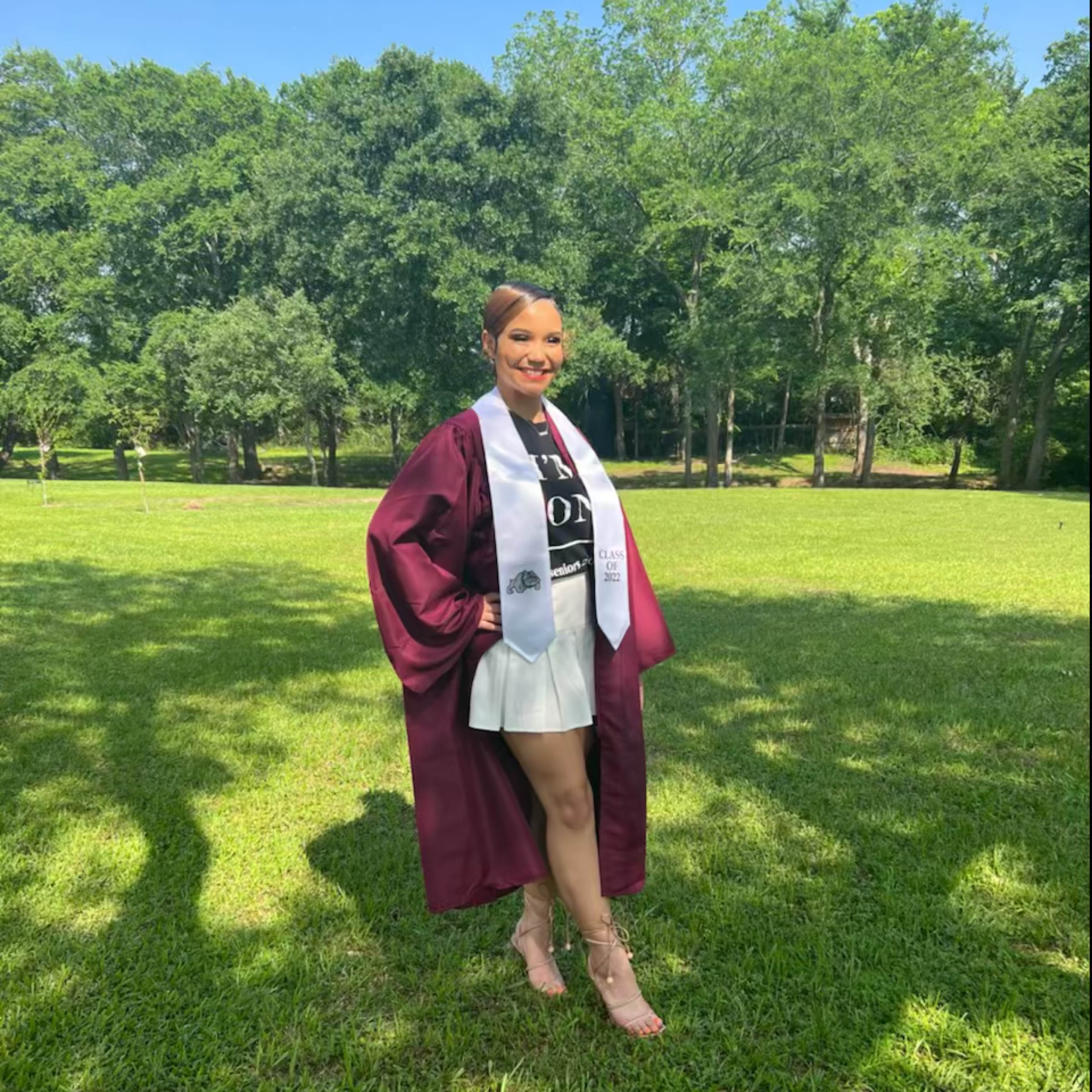 A graduate in a maroon robe and sash stands in a grassy field with trees in the background.